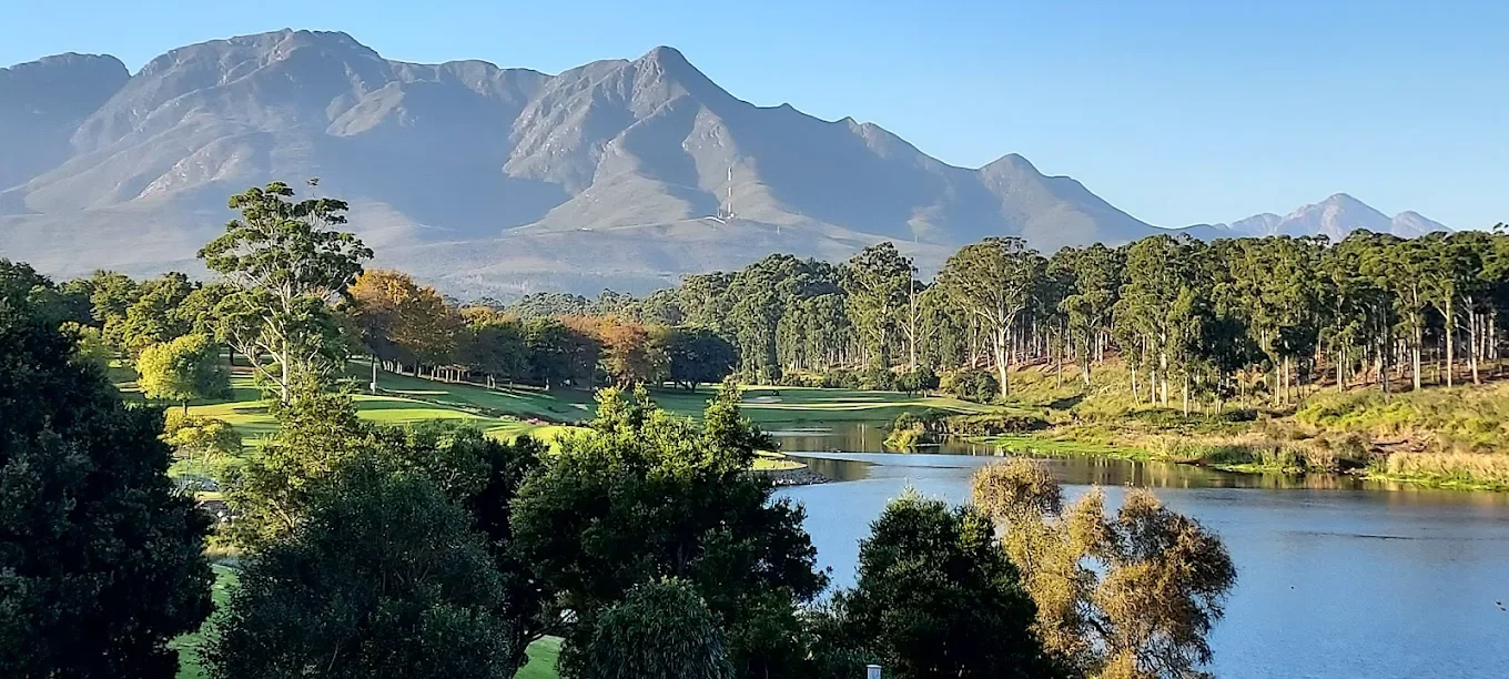 Scenic view of a river flowing through a lush green landscape with trees and a golf course, with mountains in the background under a clear blue sky.