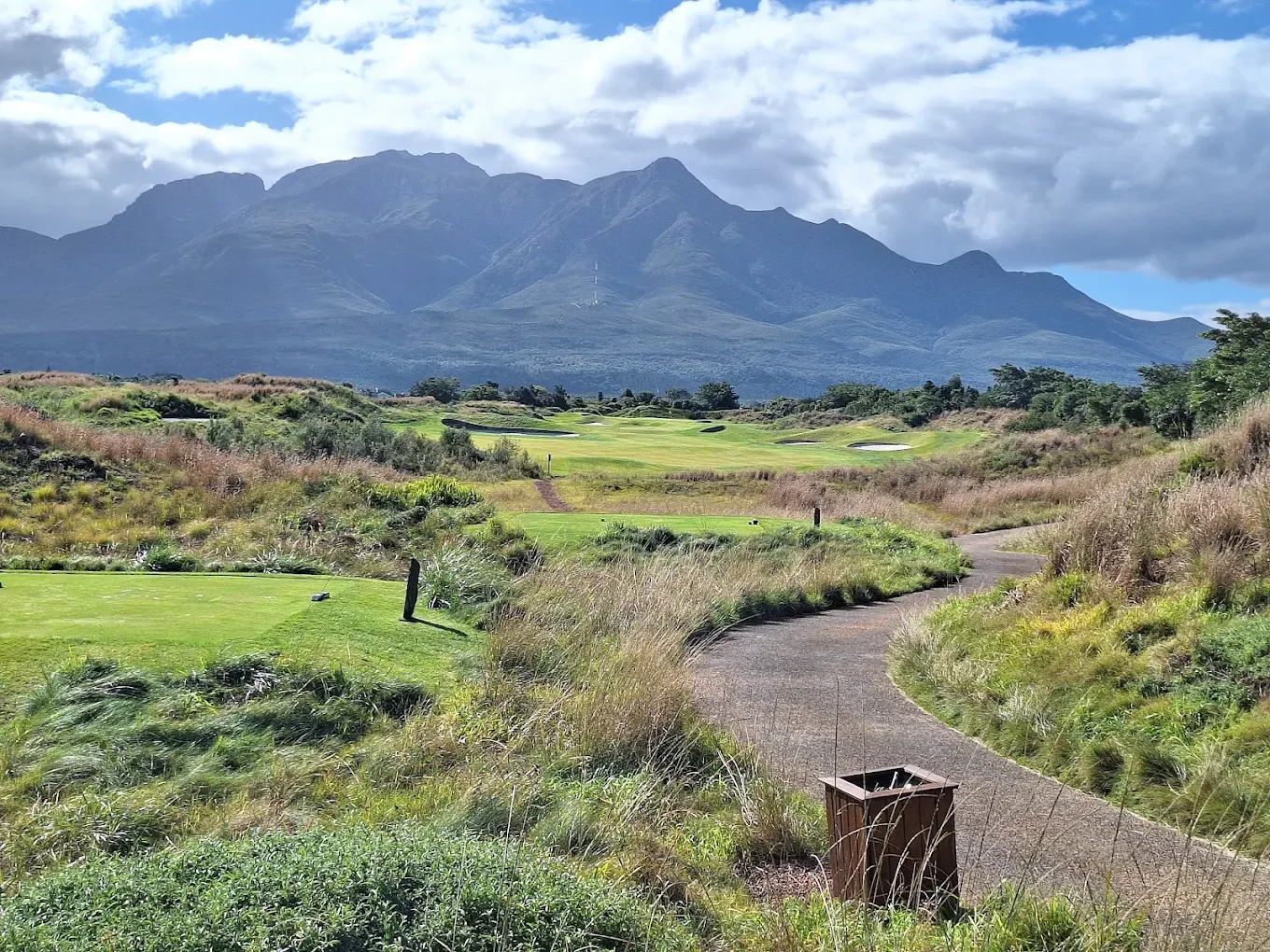 Scenic view of a golf course with a winding path, green fairways, and sand traps, set against lush grass and mountainous landscape under partly cloudy sky.