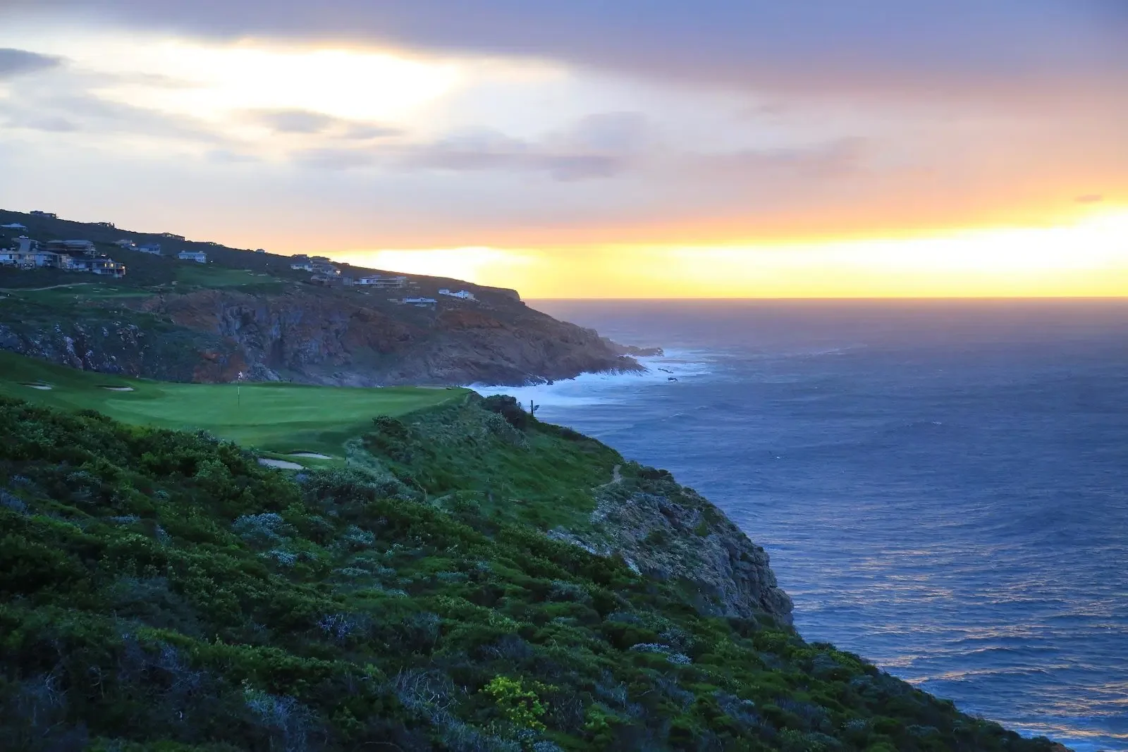 Cliffs overlooking the ocean with a golf green and houses on the hillside during sunset