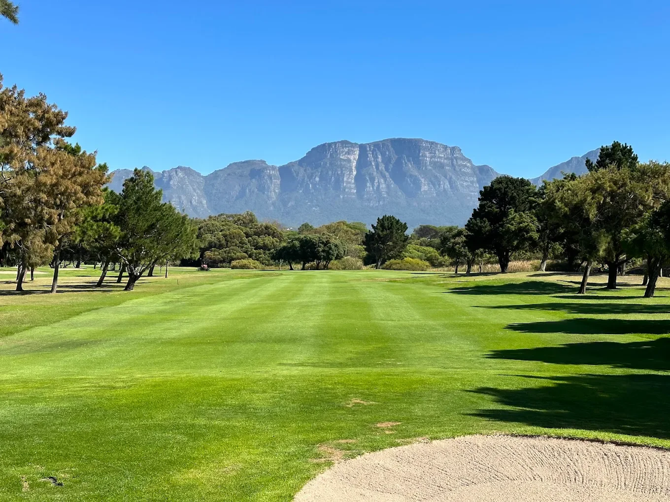 A golf course with neatly mowed green grass, surrounded by trees, with a mountain range in the background under a clear blue sky.