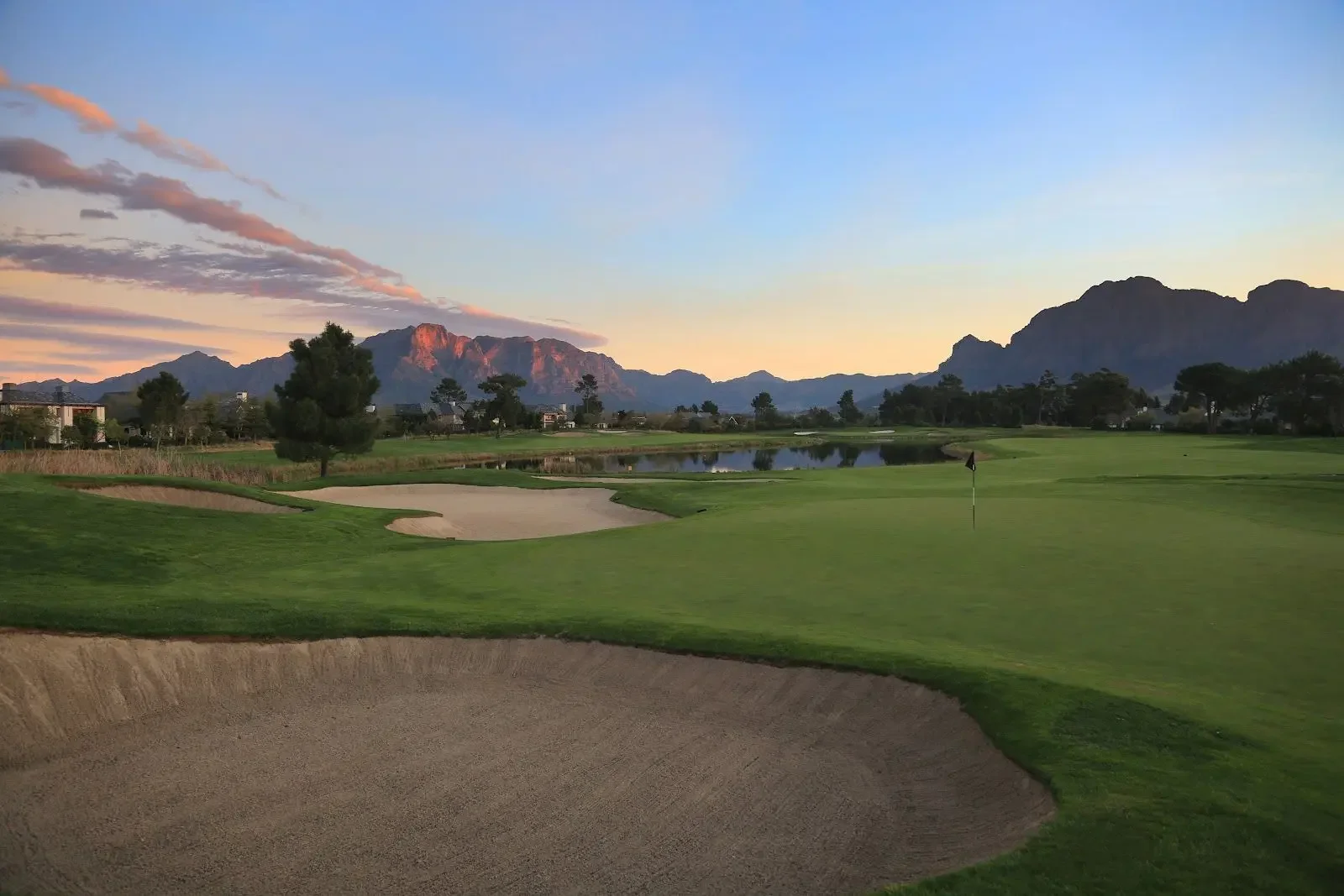 A golf course at sunset with a sand trap in the foreground and water hazard, trees, and mountains in the background.