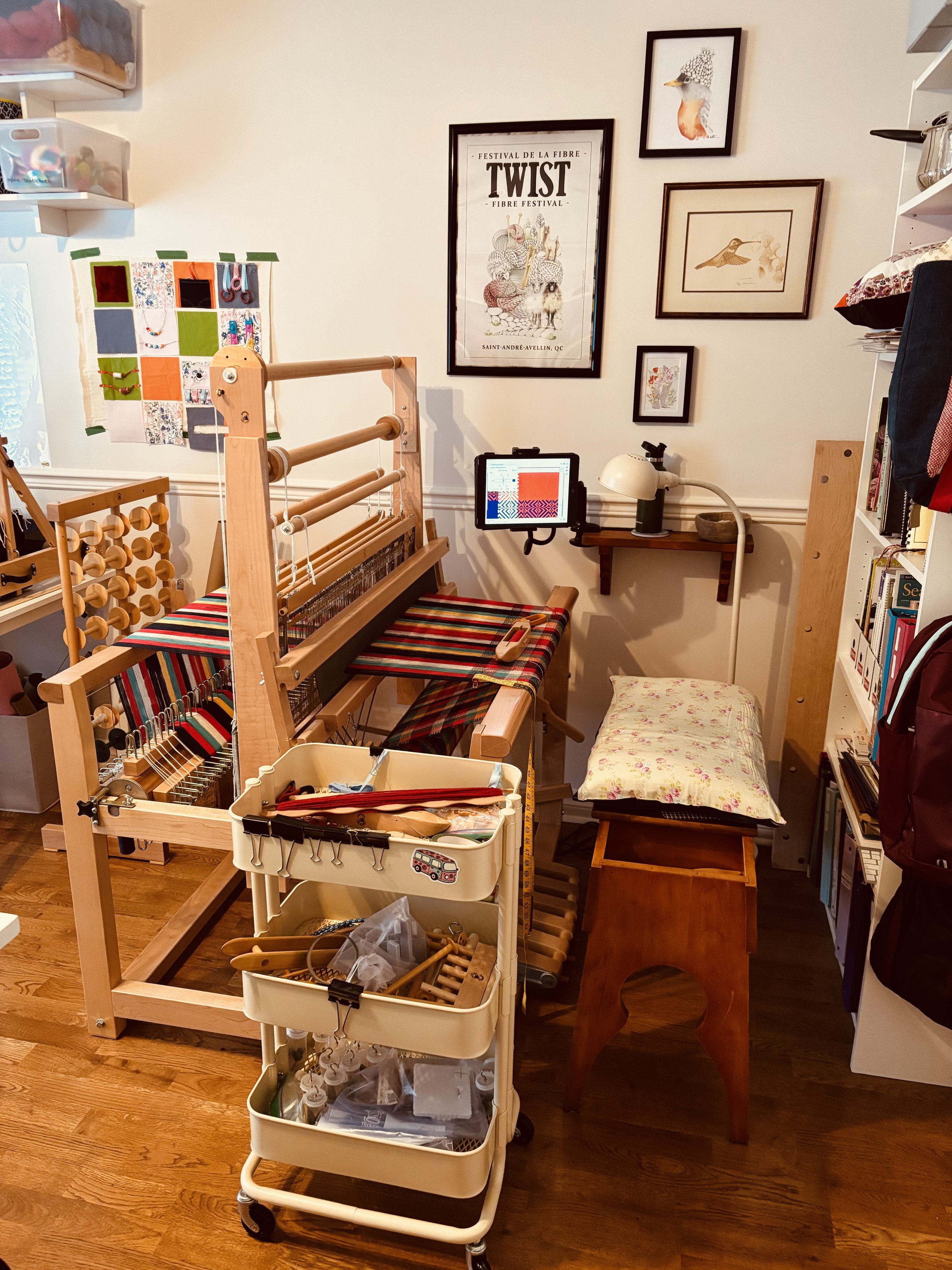 Une pièce consacrée au tissage avec un métier à tisser en bois, une petite table avec un coussin, des cadres avec des illustrations d'oiseaux et d'animaux, une tablette avec une tablette numérique, une étagère avec des livres et divers objets.