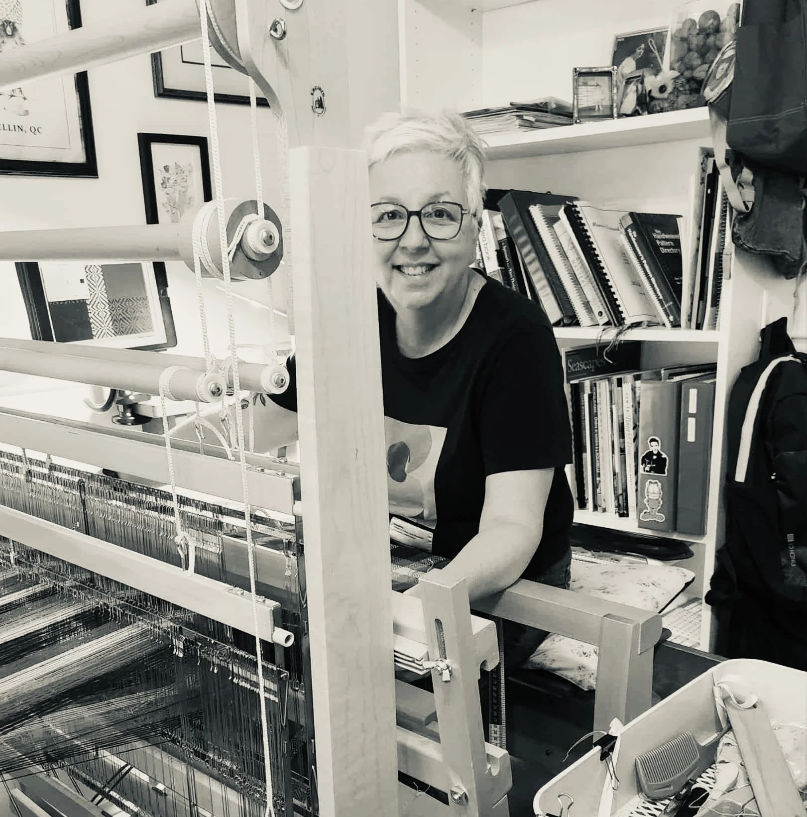 Une femme souriante avec des lunettes derrière un métier à tisser, dans une pièce remplie de livres et de décorations.