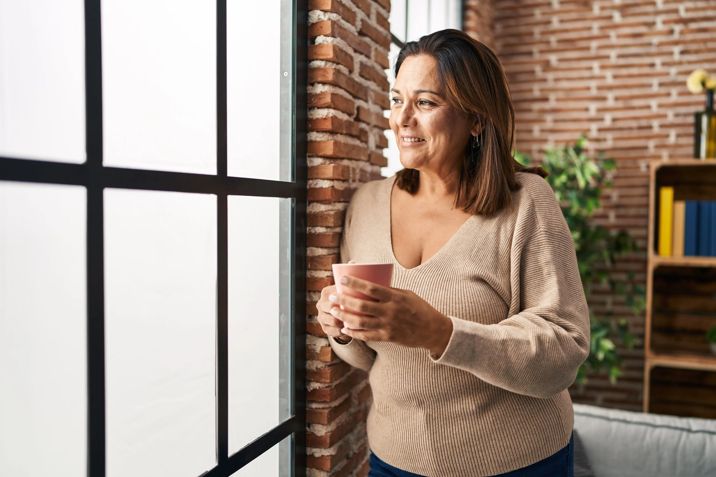 A woman with shoulder-length brown hair, wearing a beige sweater, stands by a large window holding a pink mug and smiling while looking outside. The background includes a brick wall and a bookshelf with plants and books.