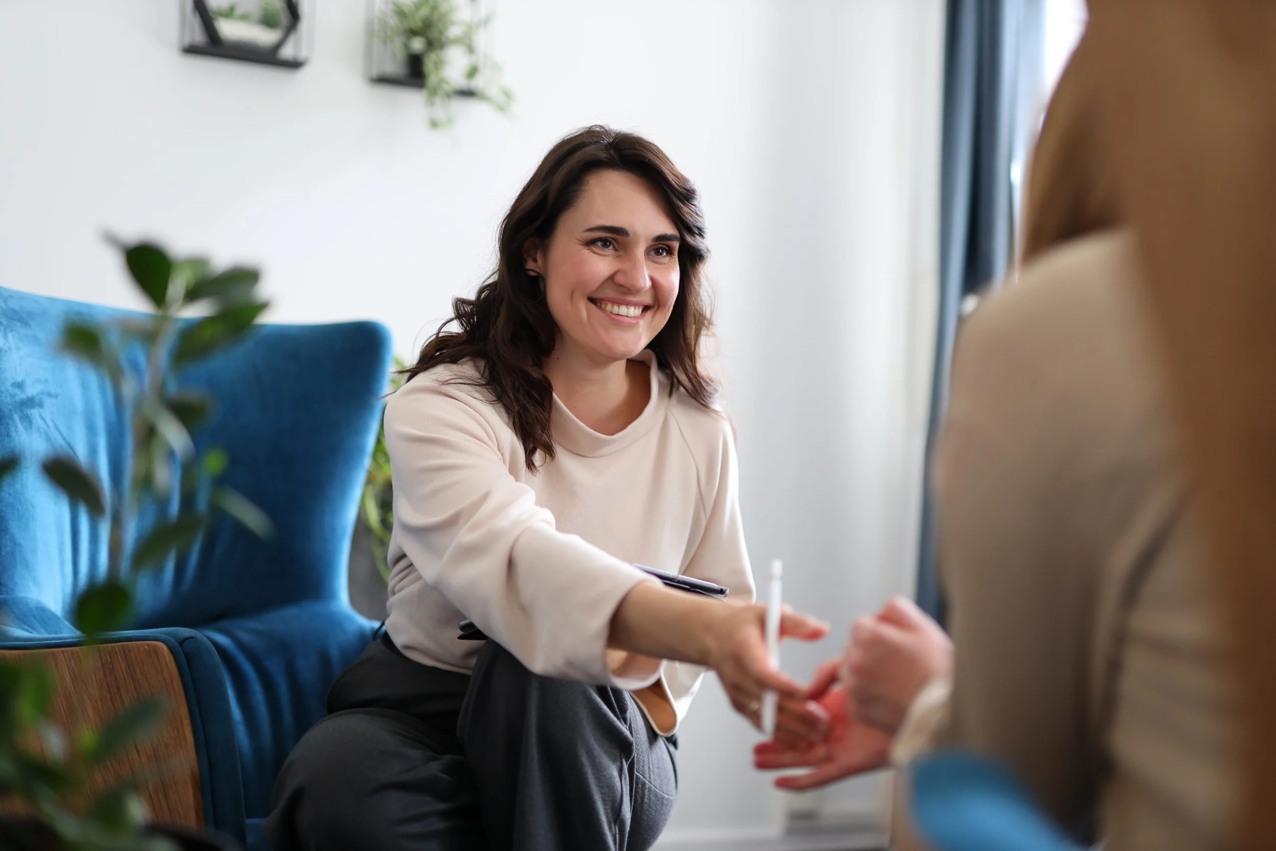 A woman smiling and shaking hands with someone, sitting on a blue chair in a bright room.
