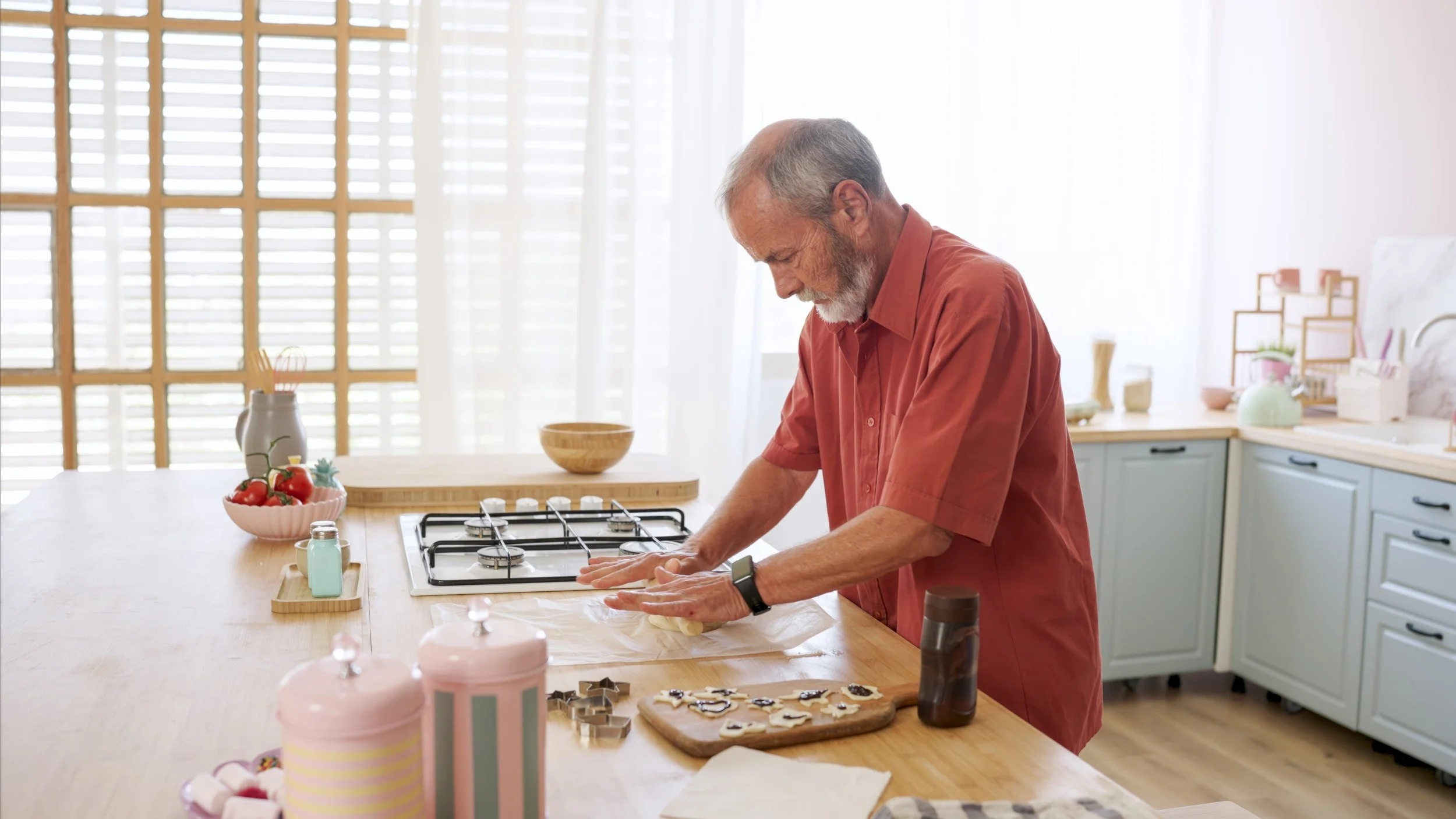 An elderly man with gray hair and beard wearing a red shirt, standing at a kitchen counter, preparing cookies with cookie cutters on a wooden board.