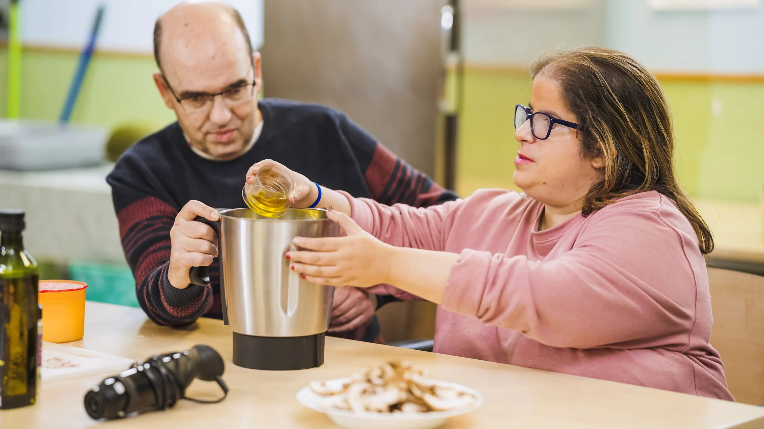 A woman with glasses and a pink sweatshirt helping a man with glasses pour olive oil into a large blender at a kitchen table.
