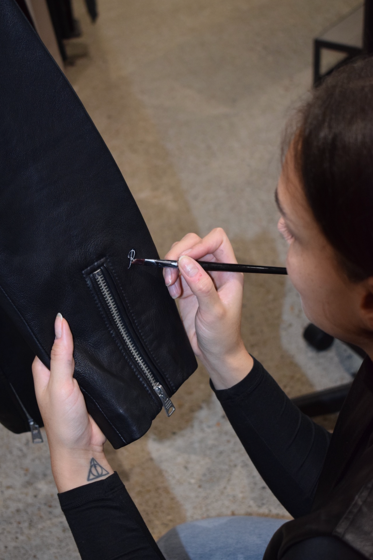 A woman with a small triangle tattoo on her wrist is sewing a small detail into a black leather bag.