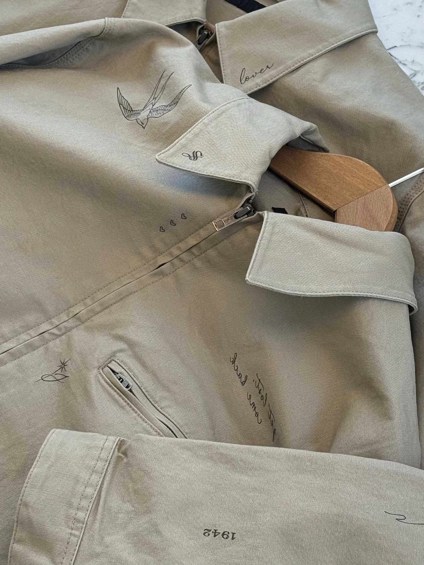 A beige jacket with embroidered designs, including a bird in flight, handwritten text, a zipper pocket, and various small symbols, laid out on a surface