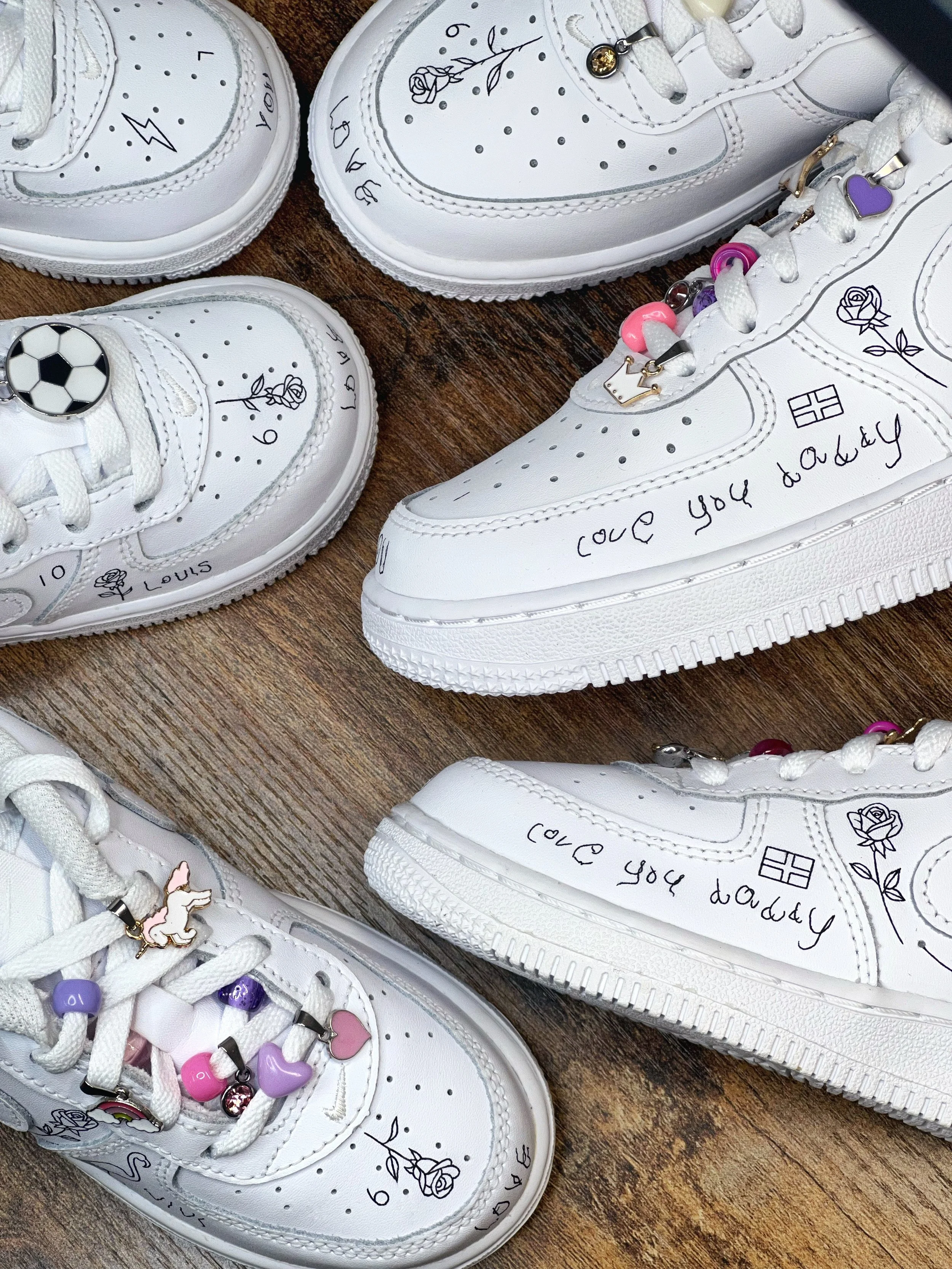 Six white sneakers decorated with drawings, charms, and handwritten messages. The sneakers feature small illustrations of roses, hearts, a soccer ball, and a flag, along with charms of a unicorn, a heart, and various beads. The handwritten messages include 'love you daddy' and other doodles.