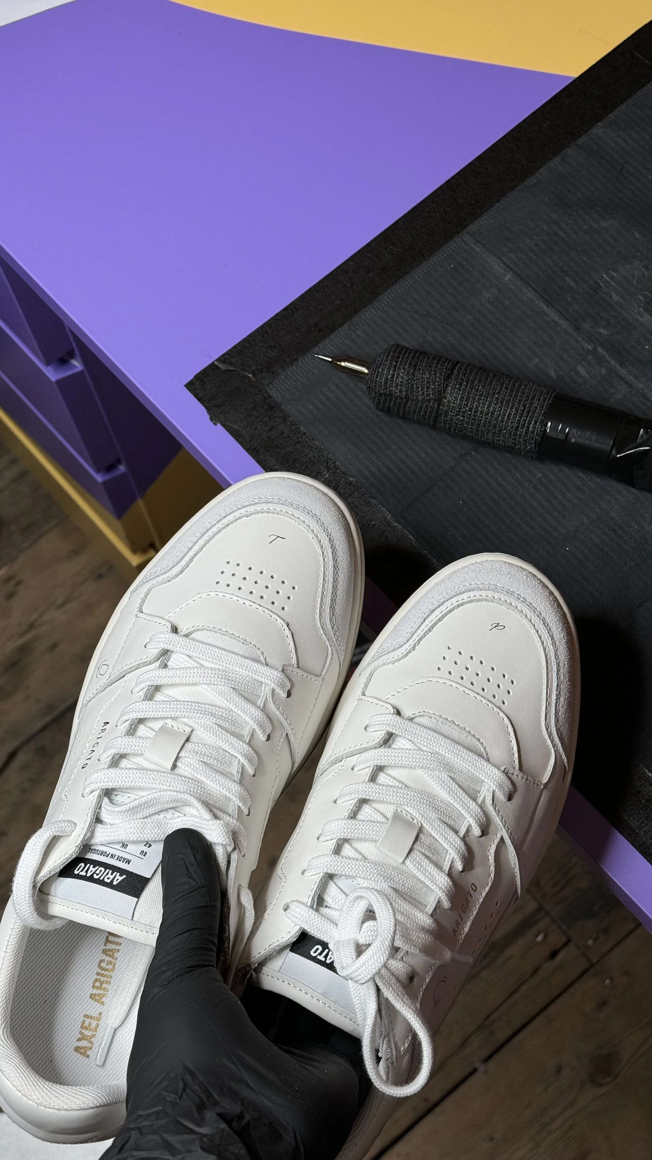A pair of white sneakers with black and gold details held by a person wearing a black glove. In the background, there is a workbench with a black tool and a purple and yellow storage unit.
