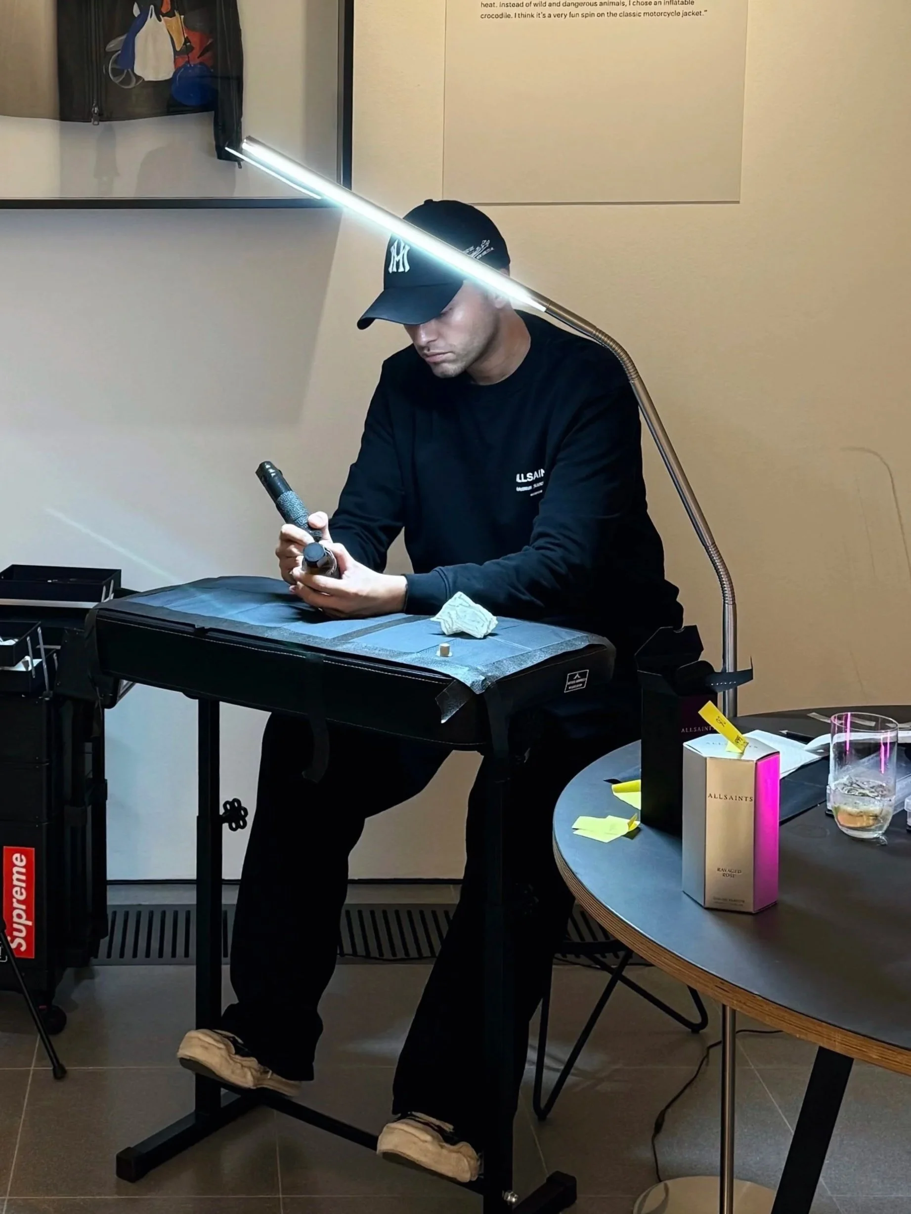A man wearing a black cap and black long-sleeve shirt sitting at a table, looking at his phone. There is a desk lamp, a box of AllSaints perfume, yellow sticky notes, and a glass with a beverage in front of him. A black Supreme sticker is visible on a cart to his left, and a mirror with a black bag hanging on it is behind him.