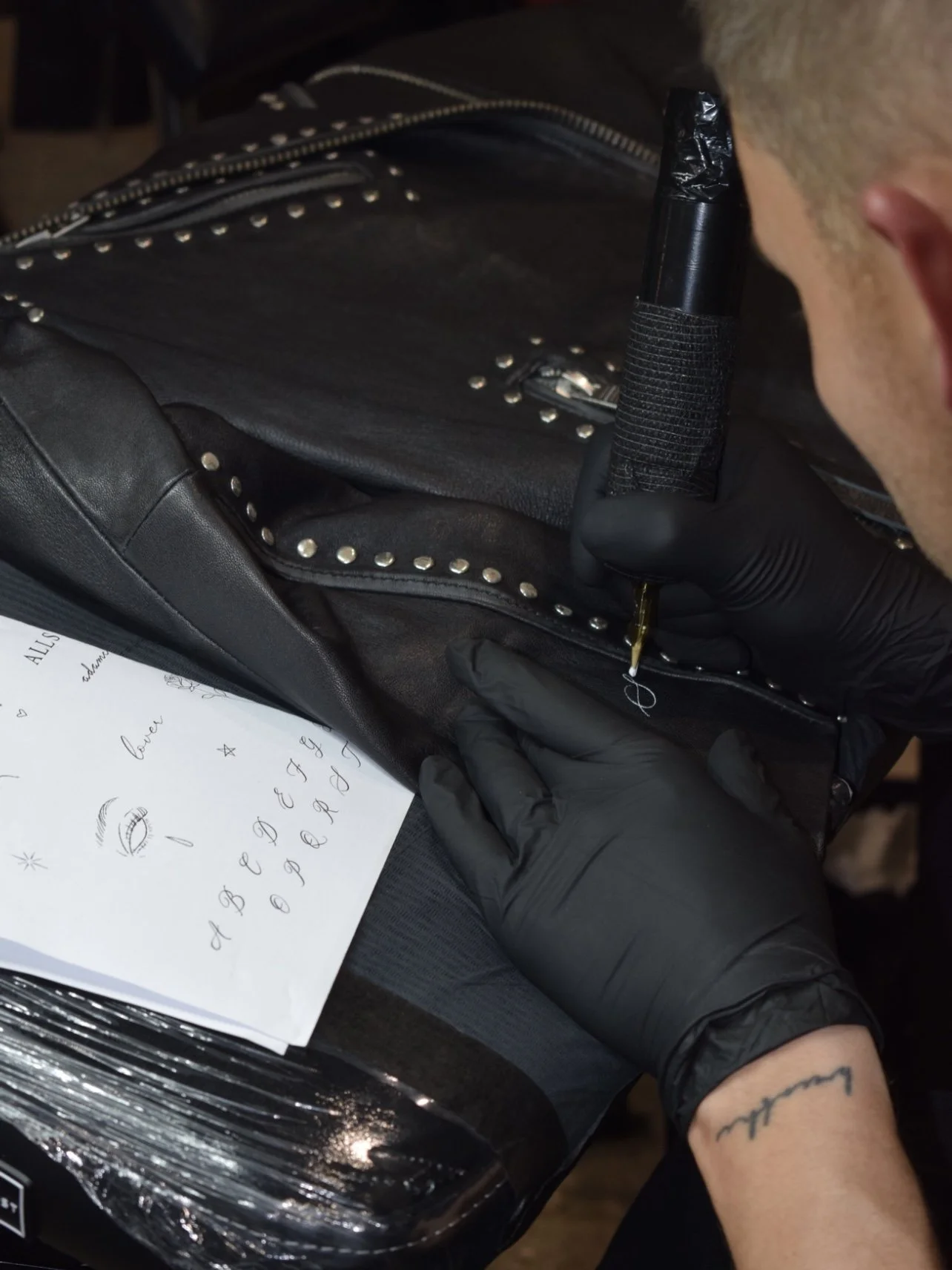 Person wearing black gloves is drawing a circle with a needle on a black leather bag that has studded accents, with a sheet of paper showing doodles and small sketches nearby.