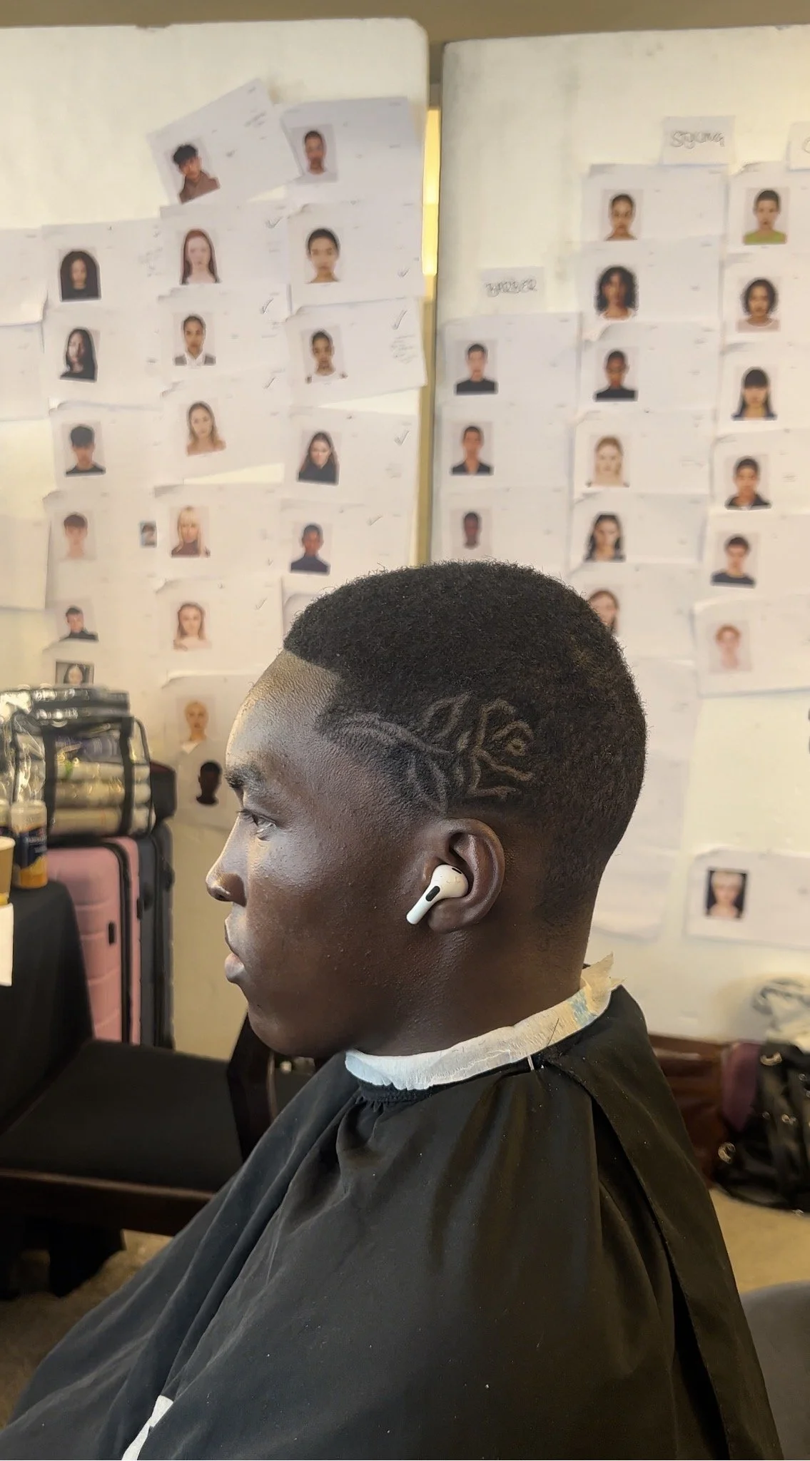 Young man with a short haircut and a decorative shaved design on the side of his head, sitting in a barber's chair with a black cape, wearing white wireless earbud, in a barbershop with photos on the wall behind him.