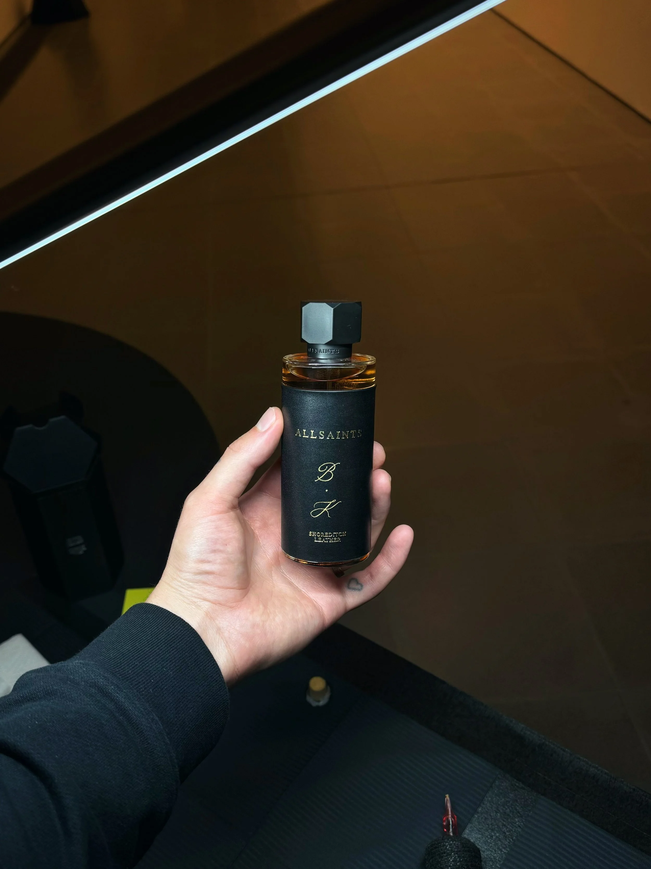 A hand holding a bottle of perfume with a black label and gold text, against a background of dark furniture.
