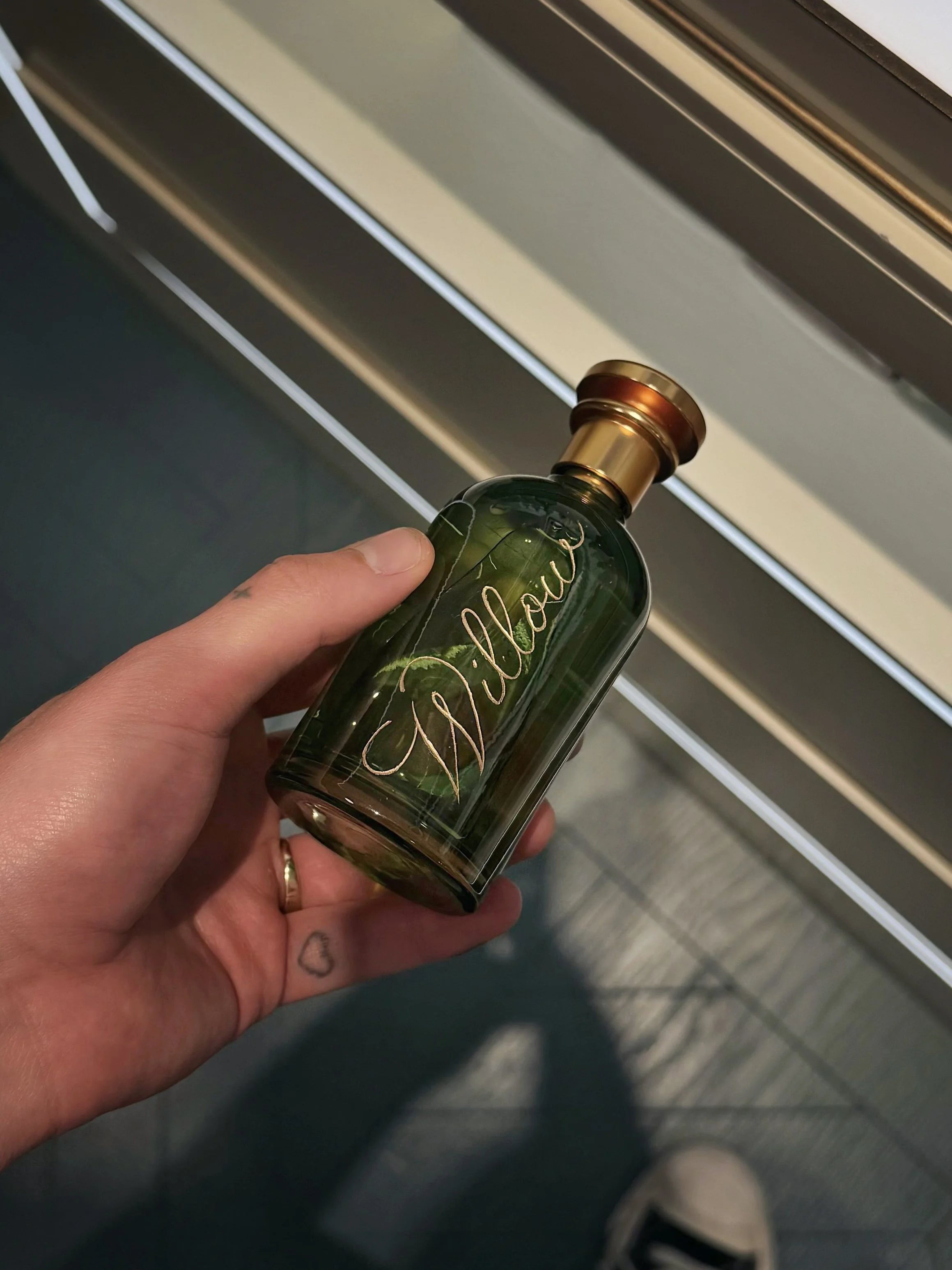 A hand holding a green glass bottle with a metallic gold cap and the name 'Willow' scripted in gold on the front, against a background of gray and beige surfaces.