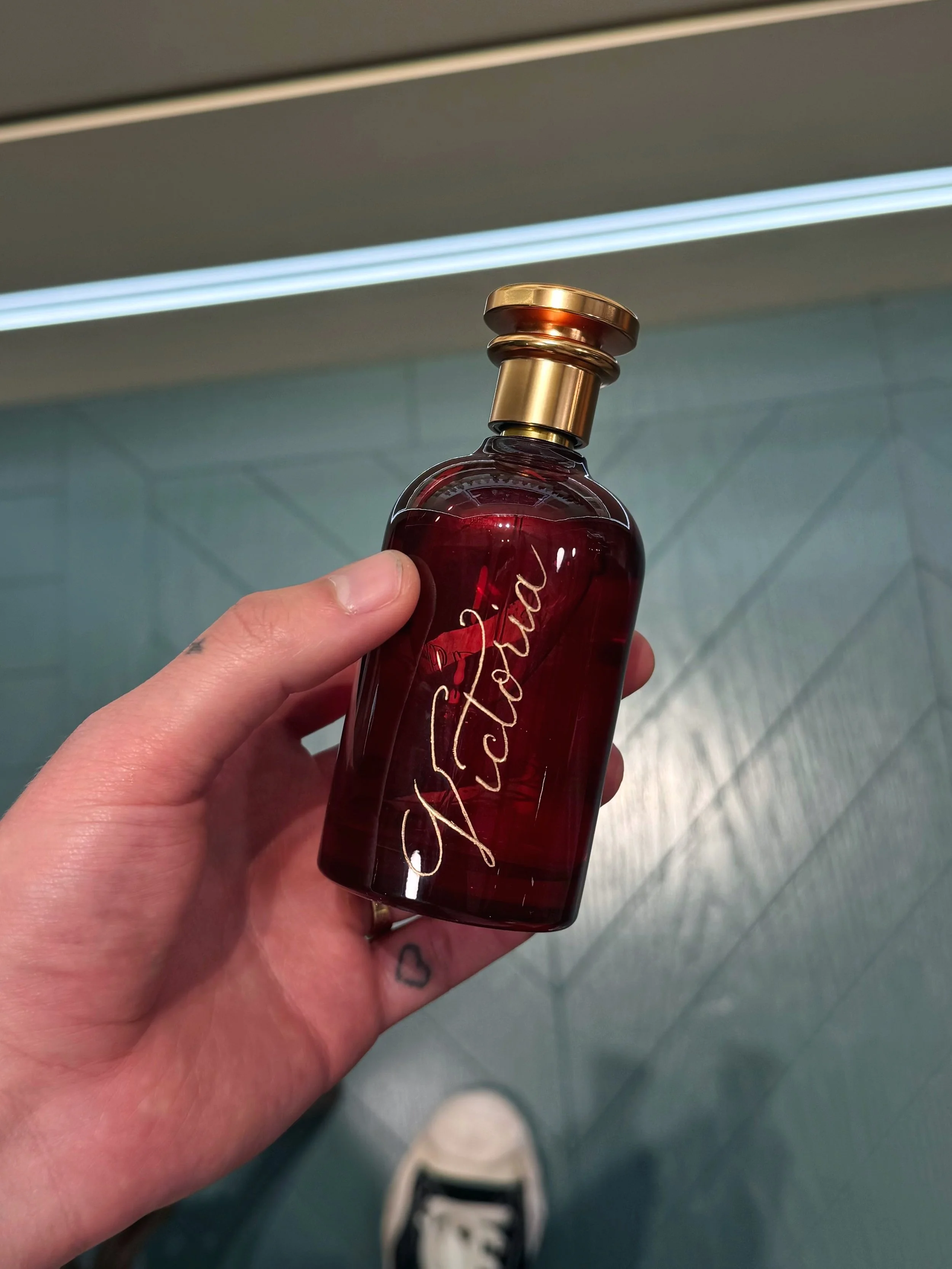 A hand holding a red perfume bottle with gold accents and cursive writing.