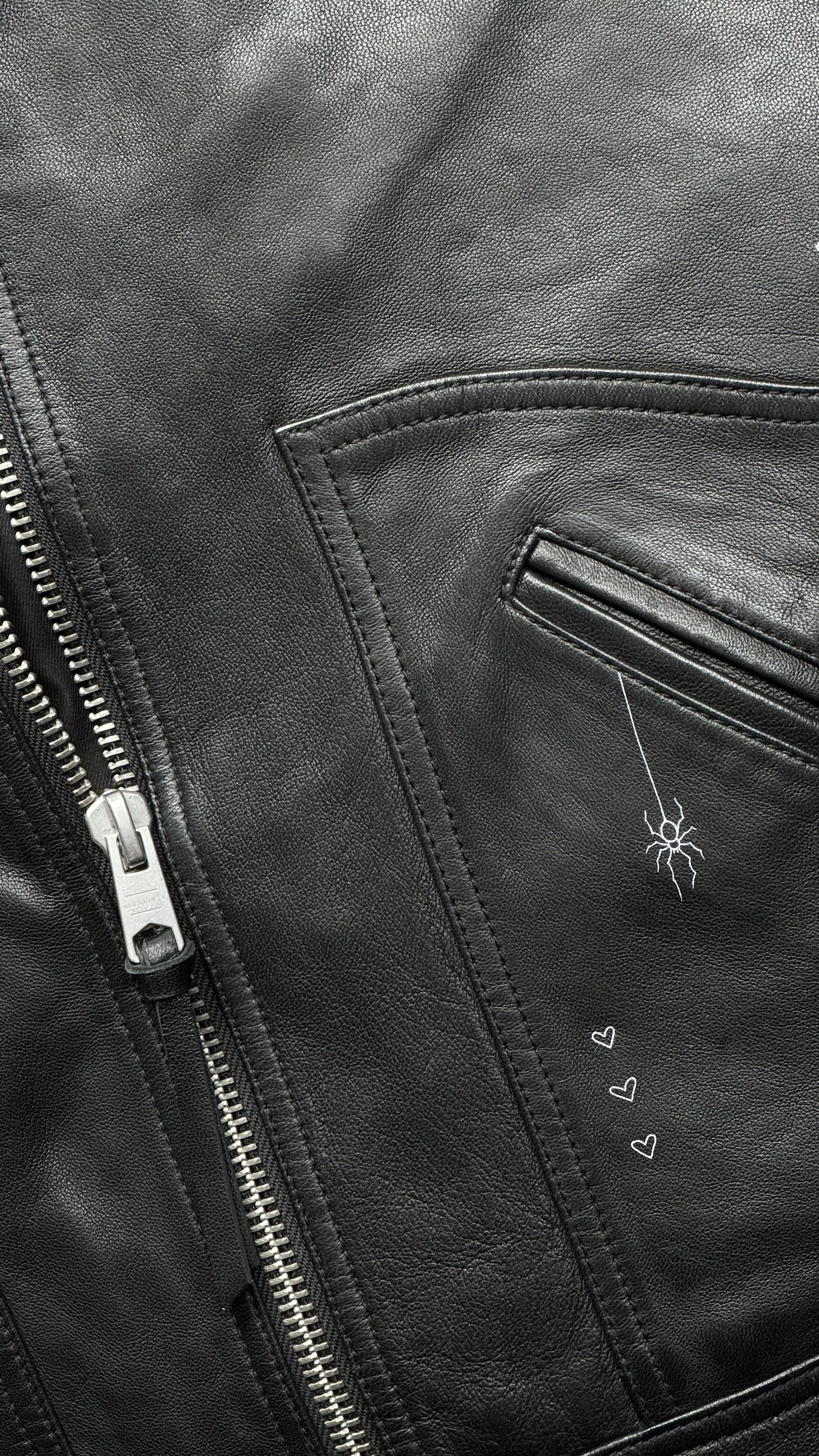 Close-up of a black leather jacket with visible stitching, a zipper on the side, and a zipper pocket. Also features a small drawn spider hanging from a web and three small hearts drawn on the leather.