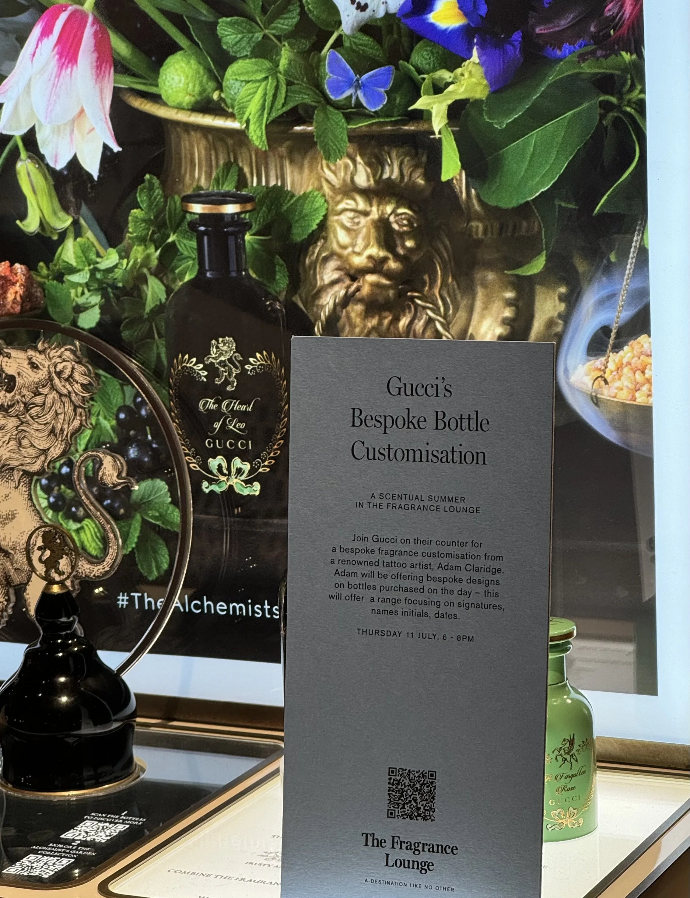 A display featuring a black Gucci fragrance bottle with gold accents, a green Gucci fragrance bottle, decorative elements including flowers, leaves, a lion's face sculpture, and a sign about Gucci's bespoke bottle customization event on July 11 from 