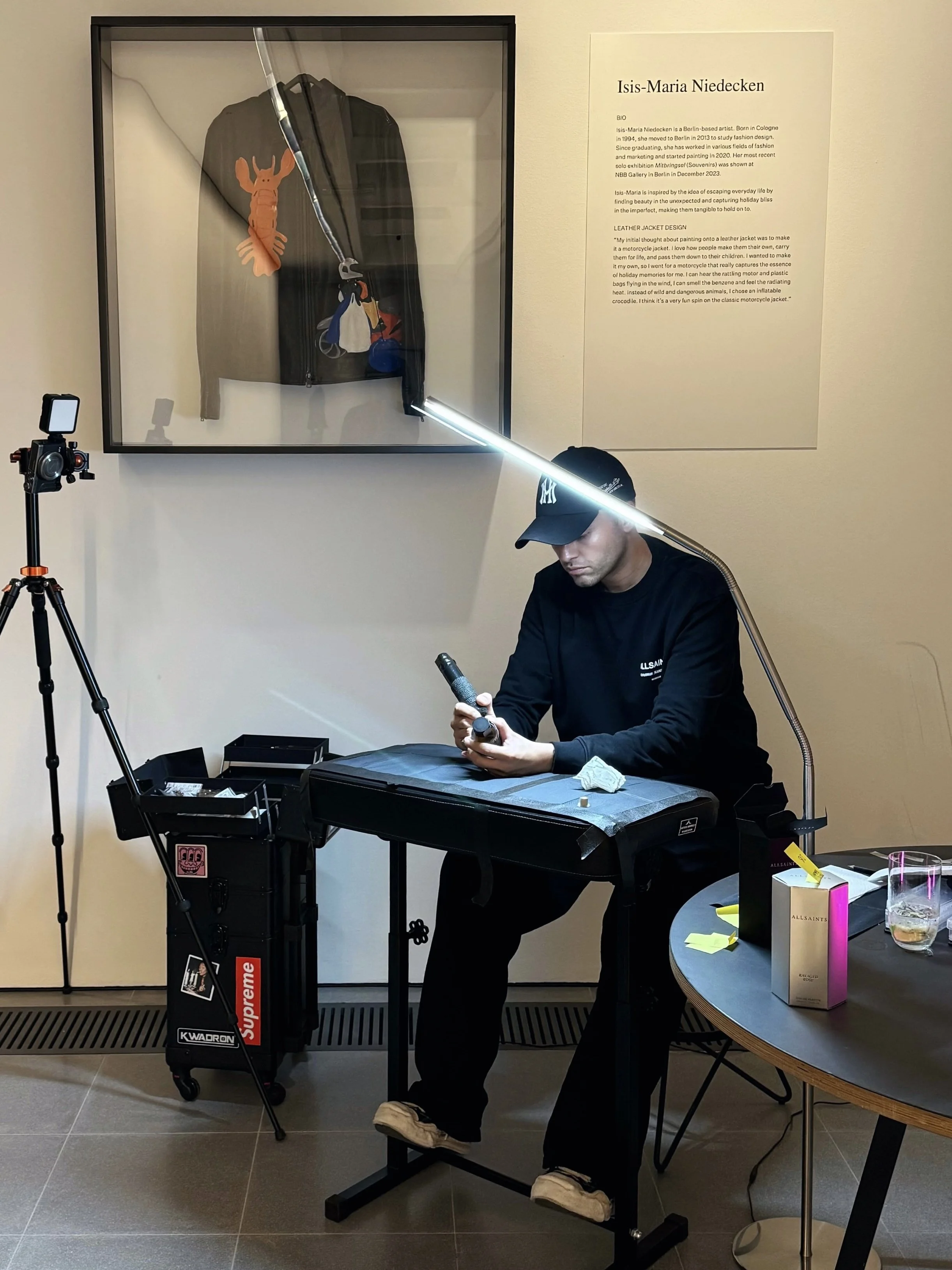 A man sitting at a table in a gallery or studio, cleaning or examining a small object with a tool. Behind him, a framed display case contains a jacket and accessories, and a wall plaque provides information about Isa-Maria Niedecken, an artist. There