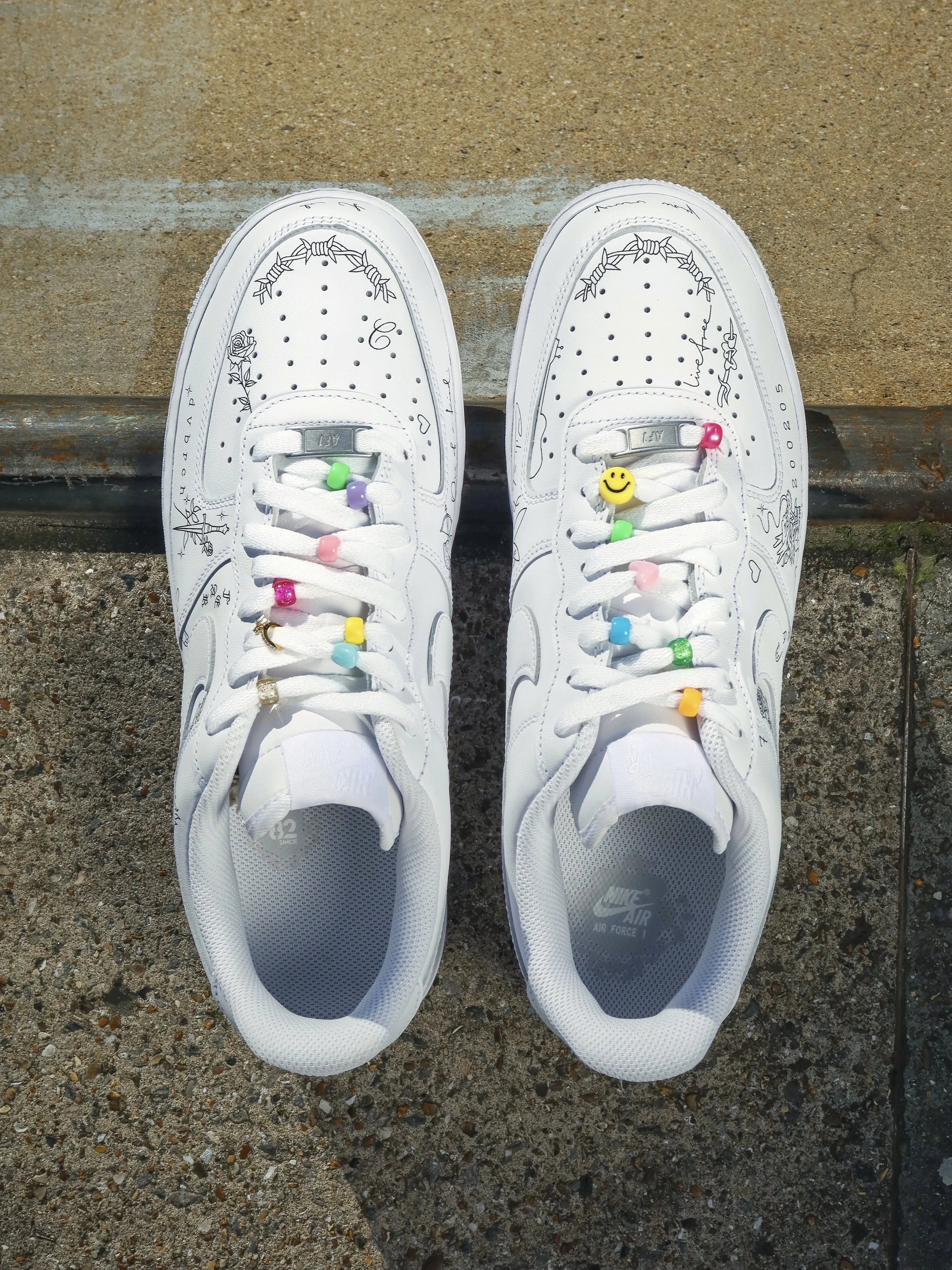 Pair of white Nike Air Force 1 sneakers with colorful beads and charms on the laces, placed on a textured concrete surface.