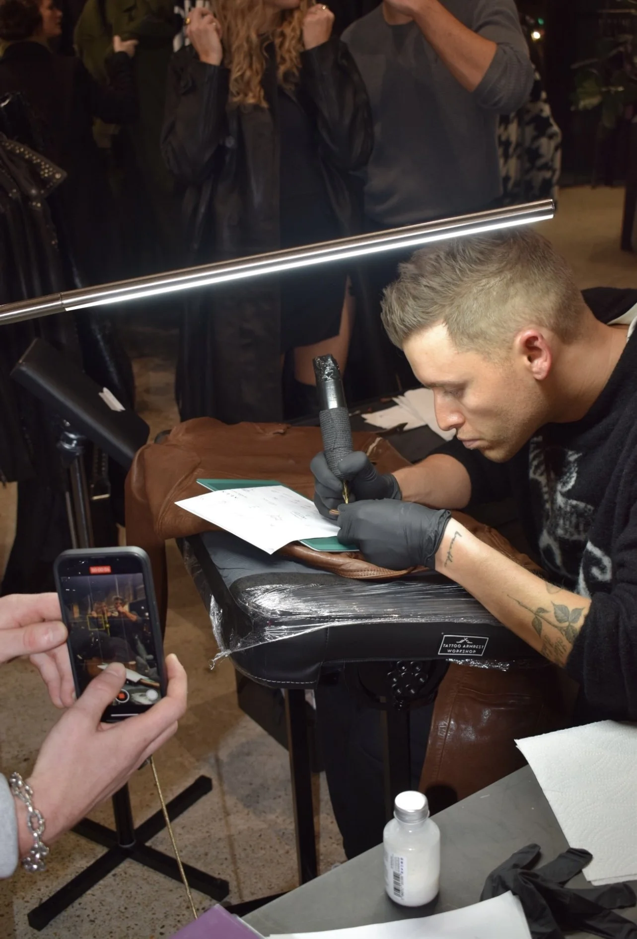A man getting a tattoo on his forearm while others watch, with a woman taking a photo on her phone.