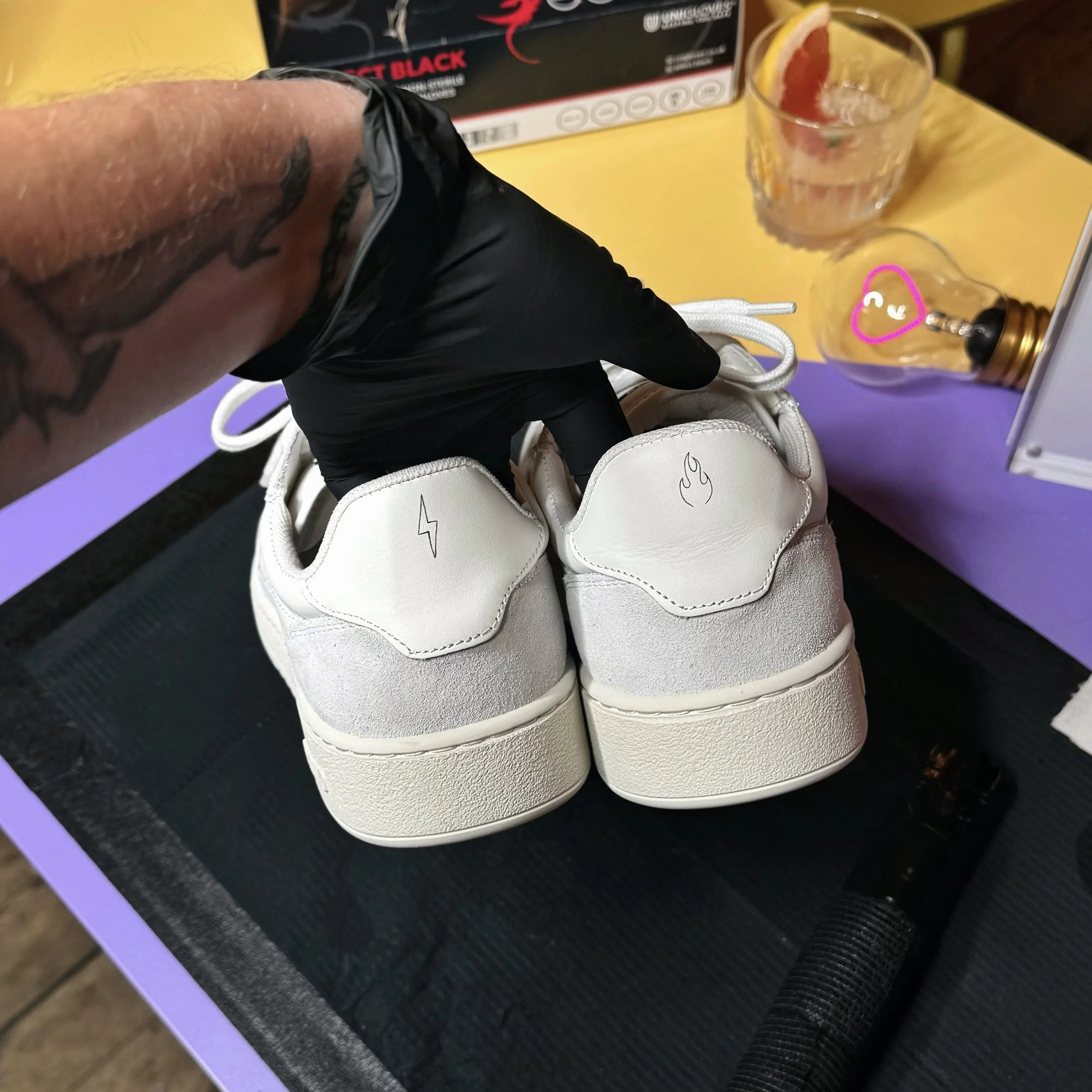 Pair of white sneakers with symbols on the heels, being held by a person wearing black gloves. In the background, there are two glasses, one with a lemon slice and the other with a heart-shaped design, on a purple surface.