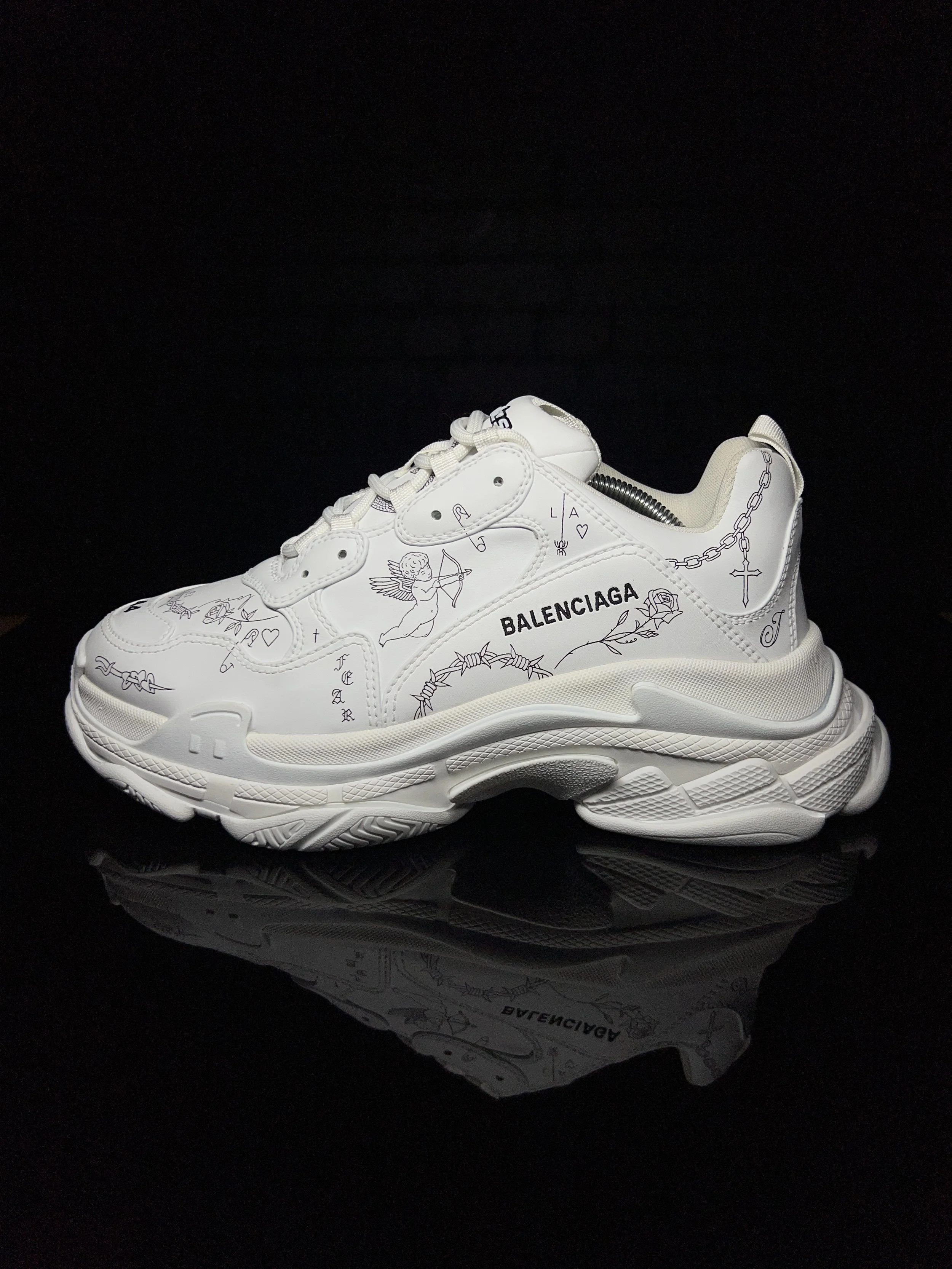 White Balenciaga sneaker with black line drawings and text, on a black reflective surface.