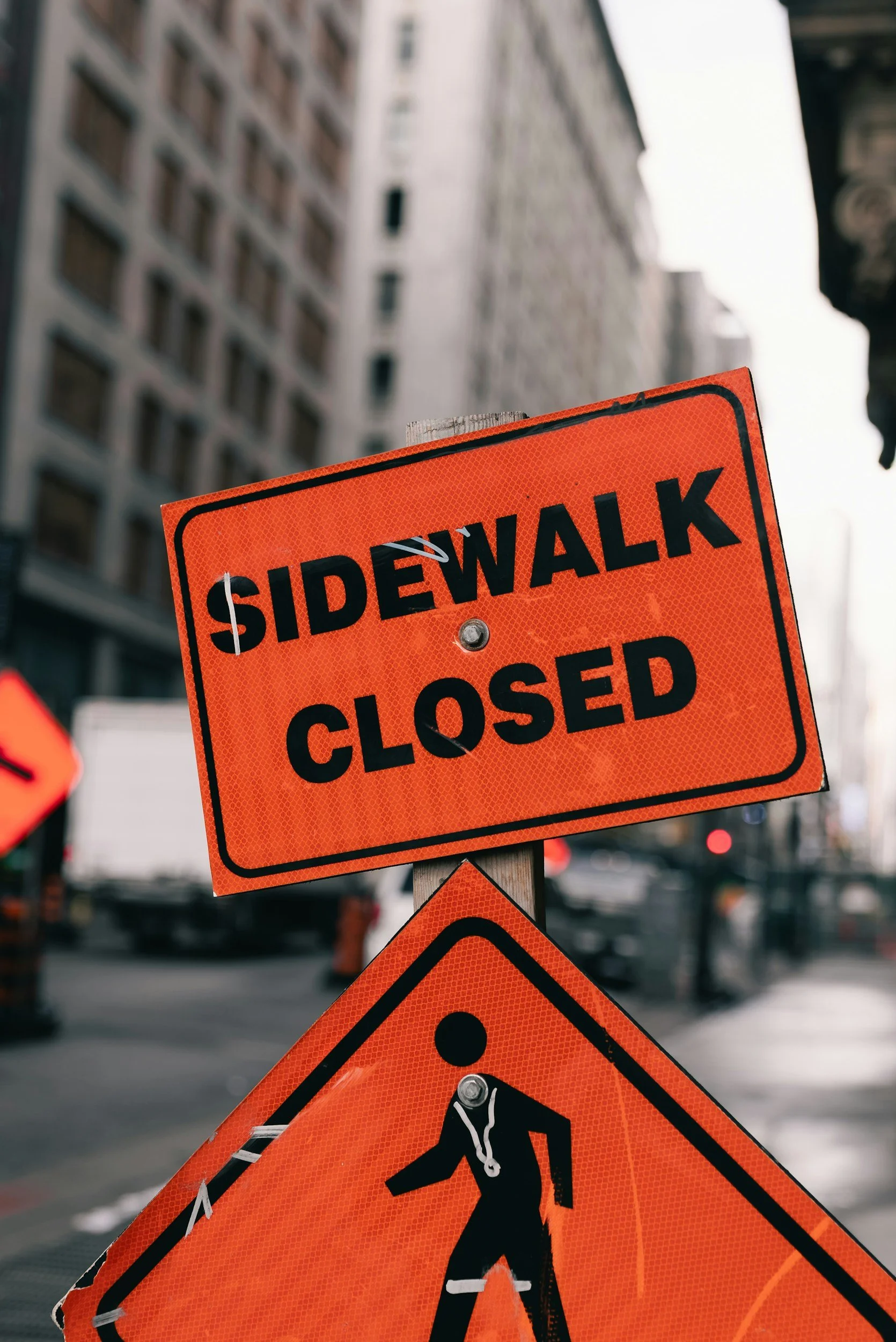 A bright orange sidewalk closed sign with black bold text, against an urban cityscape background with tall buildings and street traffic.