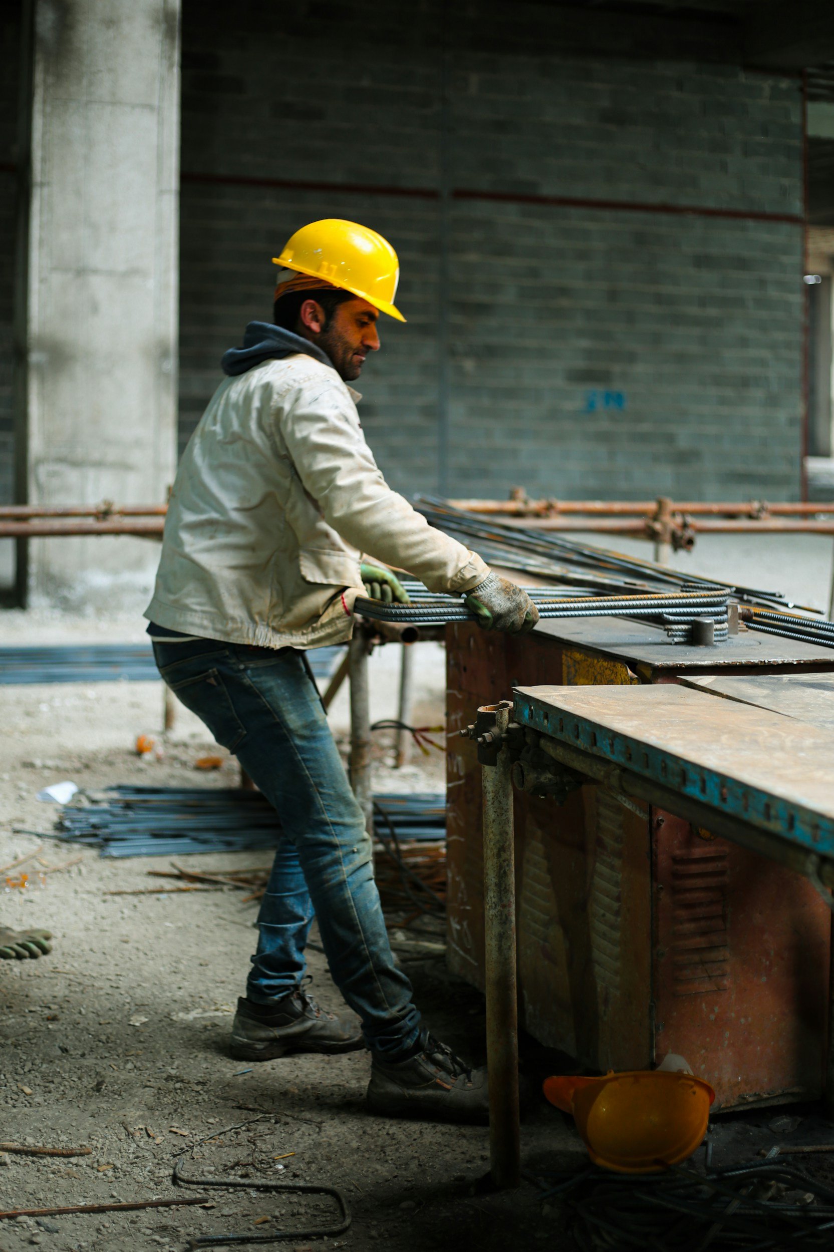 A construction worker wearing a yellow safety helmet and work gloves is working at a construction site with concrete and steel materials, with a brick wall in the background.