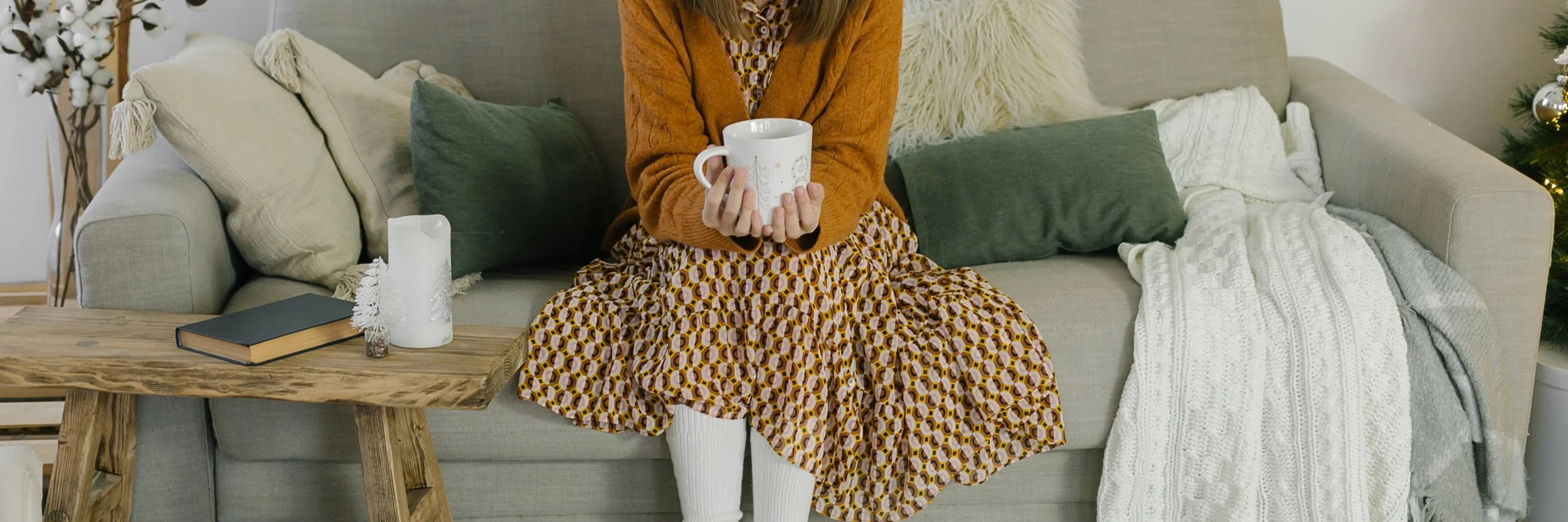 A woman sitting on a beige sofa holding a white mug, wearing a patterned dress and an orange cardigan, surrounded by cushions and Christmas decorations in a cozy living room.