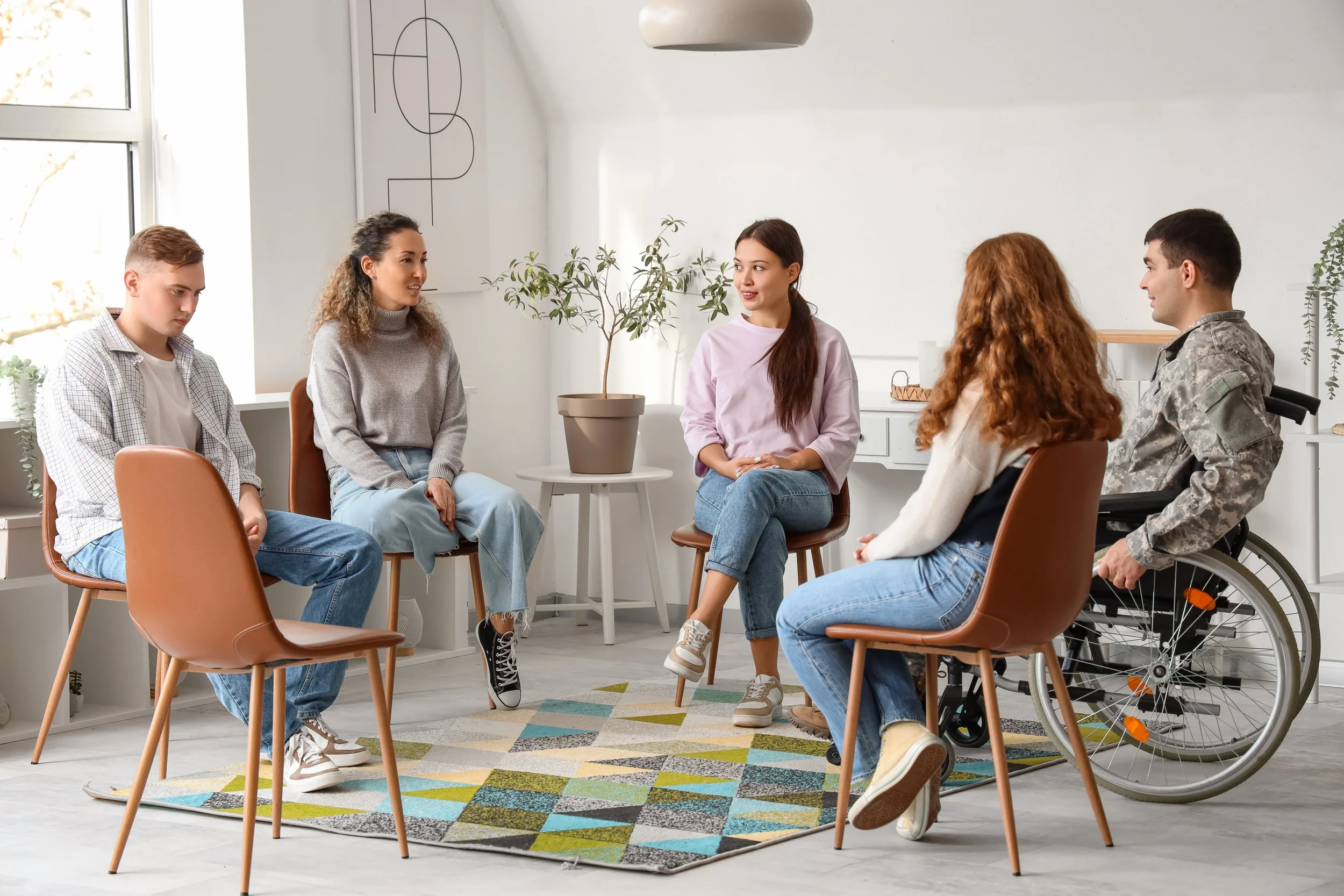 A group of six young adults in a circle having a discussion in a bright, modern room with natural light, potted plants, and abstract wall art.