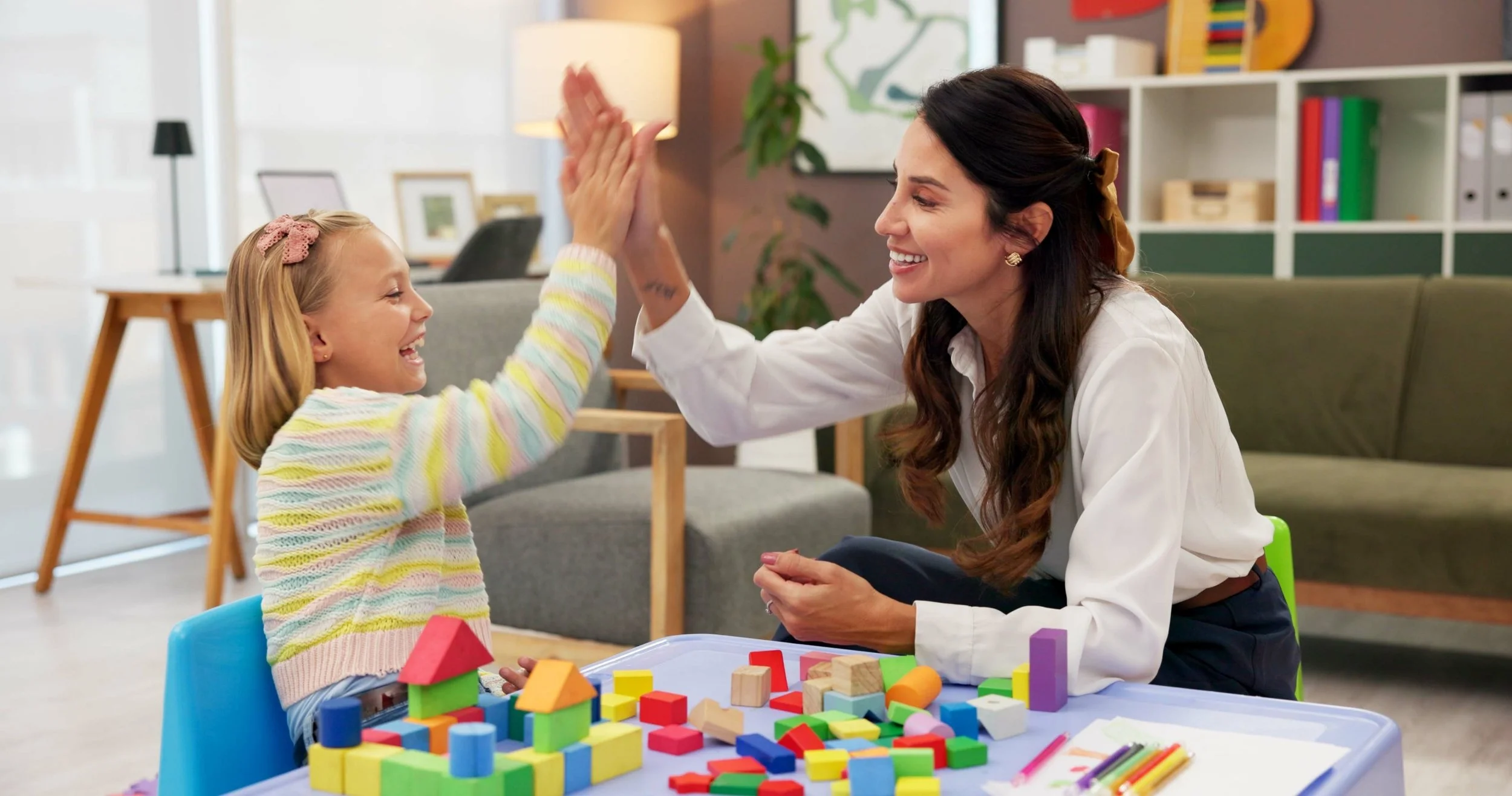 Female play therapist and child smiling and high-fiving while playing with colorful blocks at a table in a cozy room.