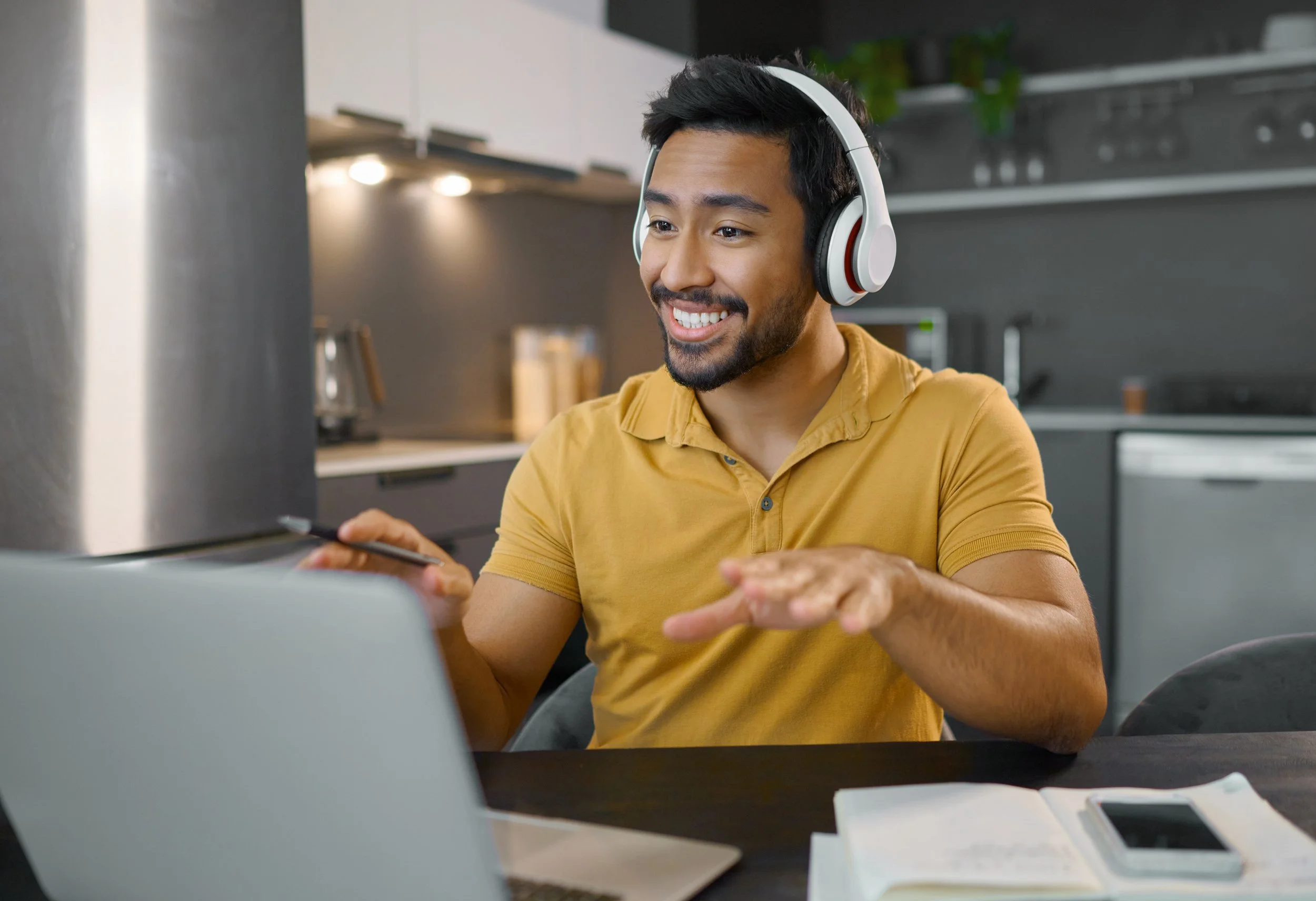 A young man smiling while wearing headphones, sitting at a table with a laptop, smartphone, and notebook in a modern kitchen.