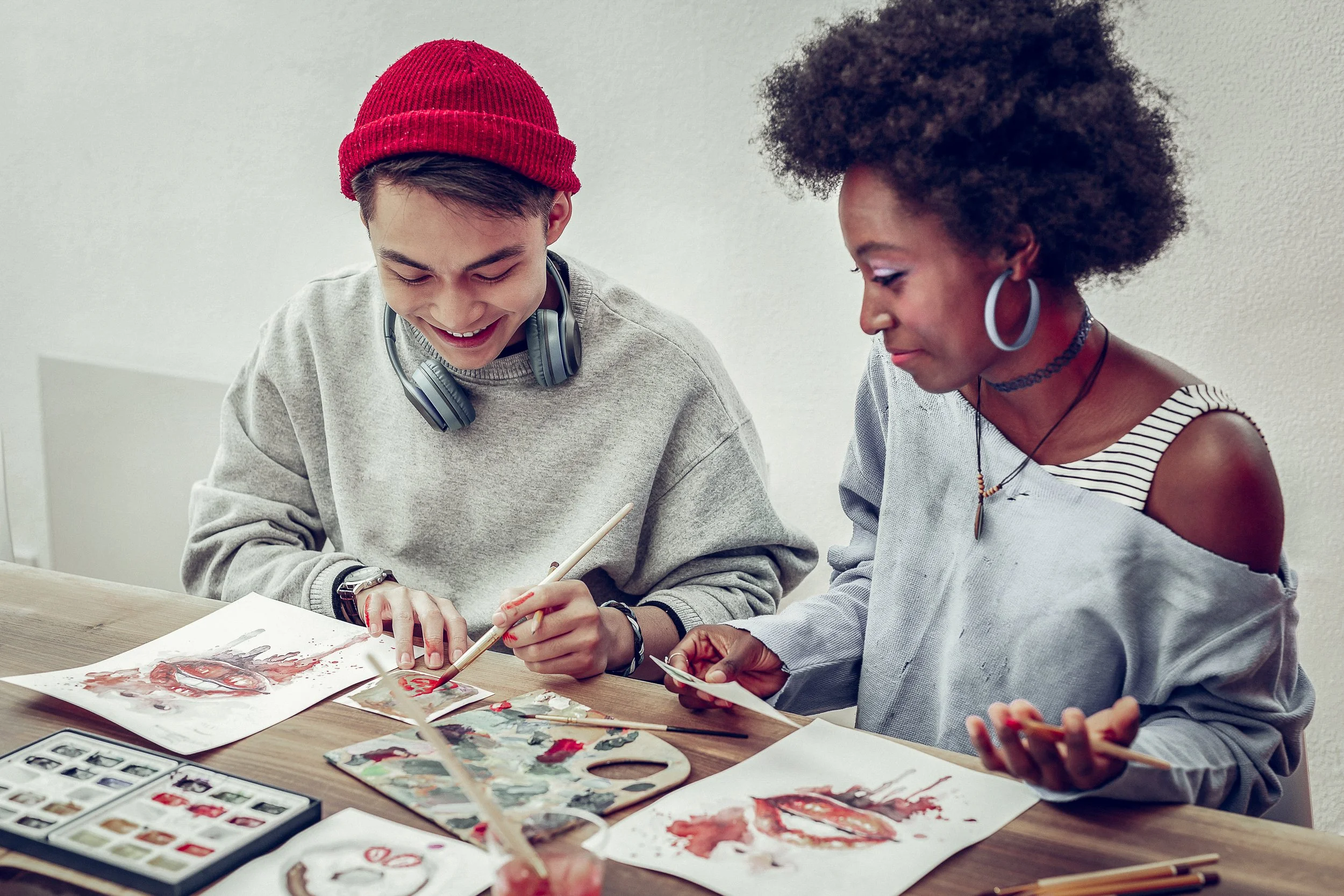 Two women sit at a table painting with watercolor. One woman is young, wearing a gray sweatshirt and red beanie, smiling as she paints. The other is older, wearing a gray and white striped shirt with cut-out shoulders, looking at her work. There are paint supplies and watercolor paintings on the table.