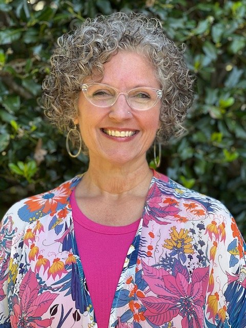 A cheerful woman with curly gray hair, wearing glasses and gold hoop earrings, stands outdoors in front of green foliage. She is dressed in a colorful floral jacket over a bright pink shirt.