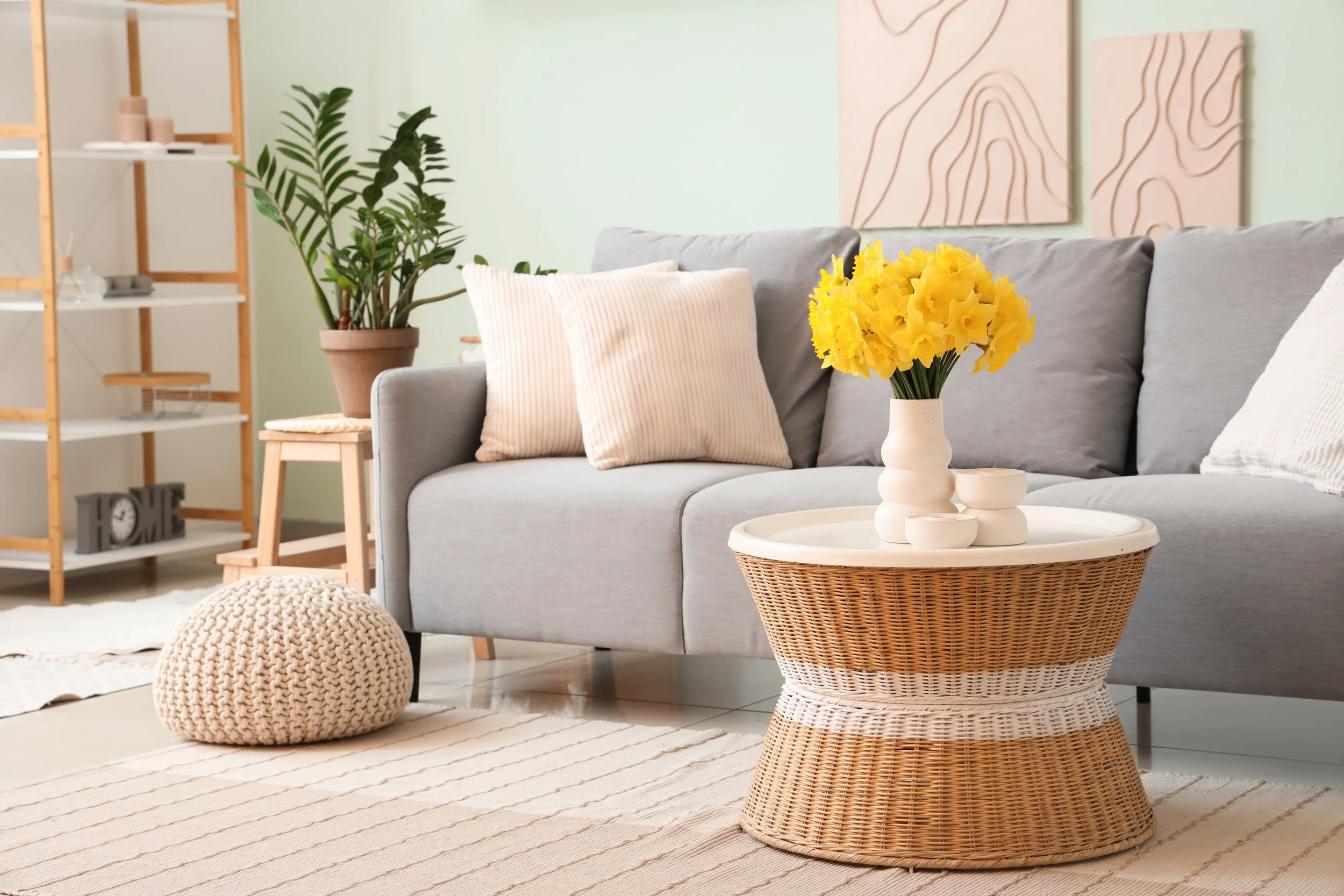 A cozy living room with a gray sofa, cream and striped pillows, a round woven coffee table with a white top, a white vase with yellow flowers, a knitted pouf, a potted plant, a cream-colored rug, and a bookshelf with decorative items and wall art with abstract line drawings.