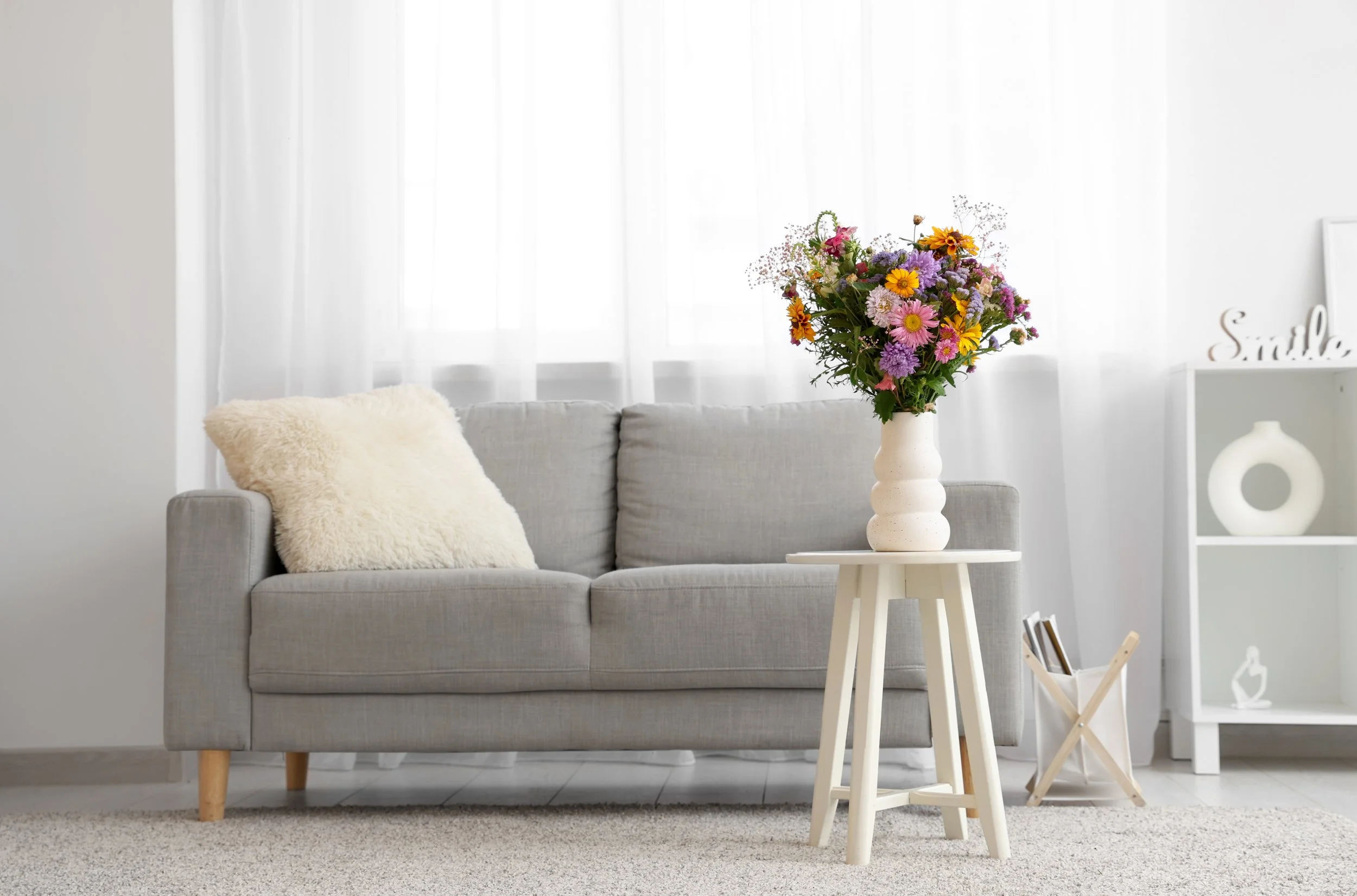 A living room with a gray sofa, a white fluffy pillow, a white side table with a vase of colorful flowers, and white shelves with decorative items in a bright, minimalistic style.