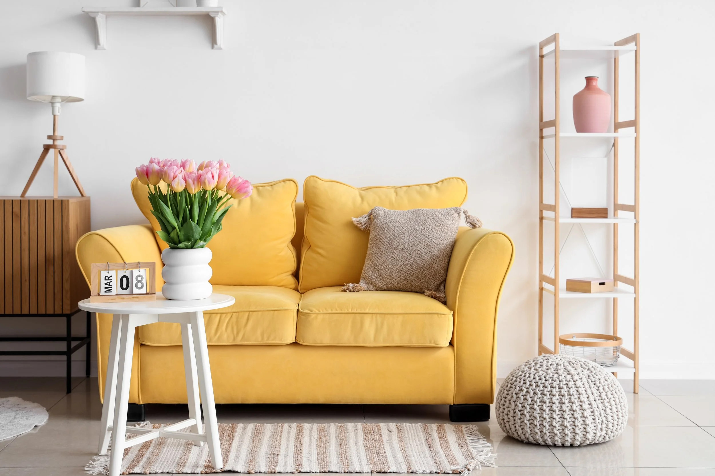 A living room with a yellow couch, beige pillow, a small white side table with pink tulips, a wooden lamp, a wooden bookshelf with a pink vase and decorative items, a knitted pouf, and a striped rug.