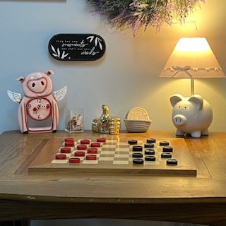 A wooden table with a chessboard and pieces, two pig-themed coin banks (one pink with wings and one white), a small gold decorative animal, a basket, and a table lamp with a shade. A chalkboard with the number 70 and some lettering hangs on the wall above.