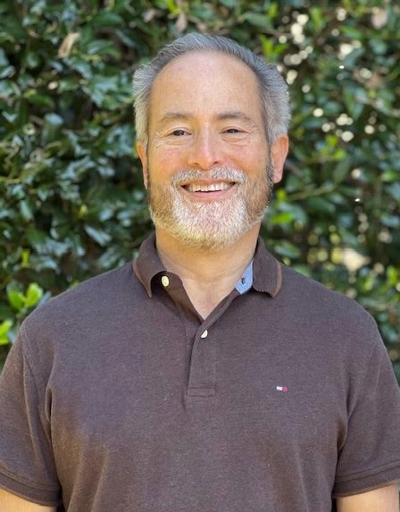 A smiling middle-aged man with gray hair and a beard, wearing a dark polo shirt, standing outdoors with green foliage in the background.