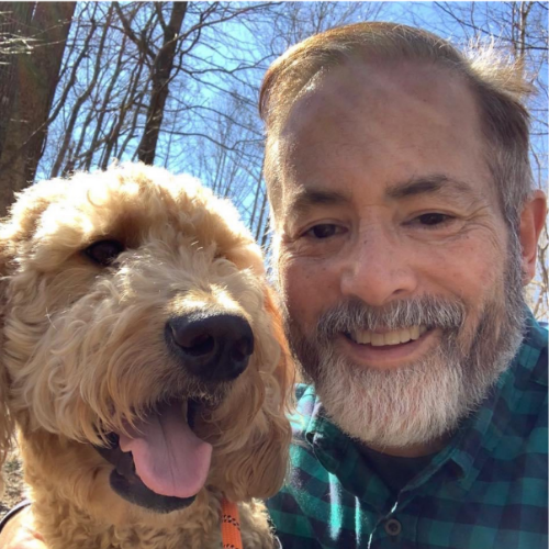 A man smiling next to a happy, curly-haired dog outdoors on a sunny day with trees in the background.
