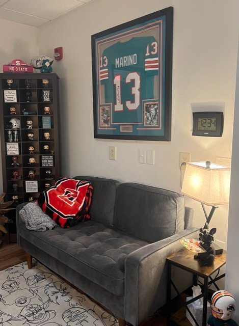 A living room corner featuring a gray velvet couch with a North Carolina State University blanket and a sweater on it. Above the couch, a large framed sports jersey of number 13 with the name Marino hangs on the wall. To the right, a wall clock shows 12:34, and a table lamp is turned on. To the left, a black display case with collectible items, and on top, a red NC State hat and figure.