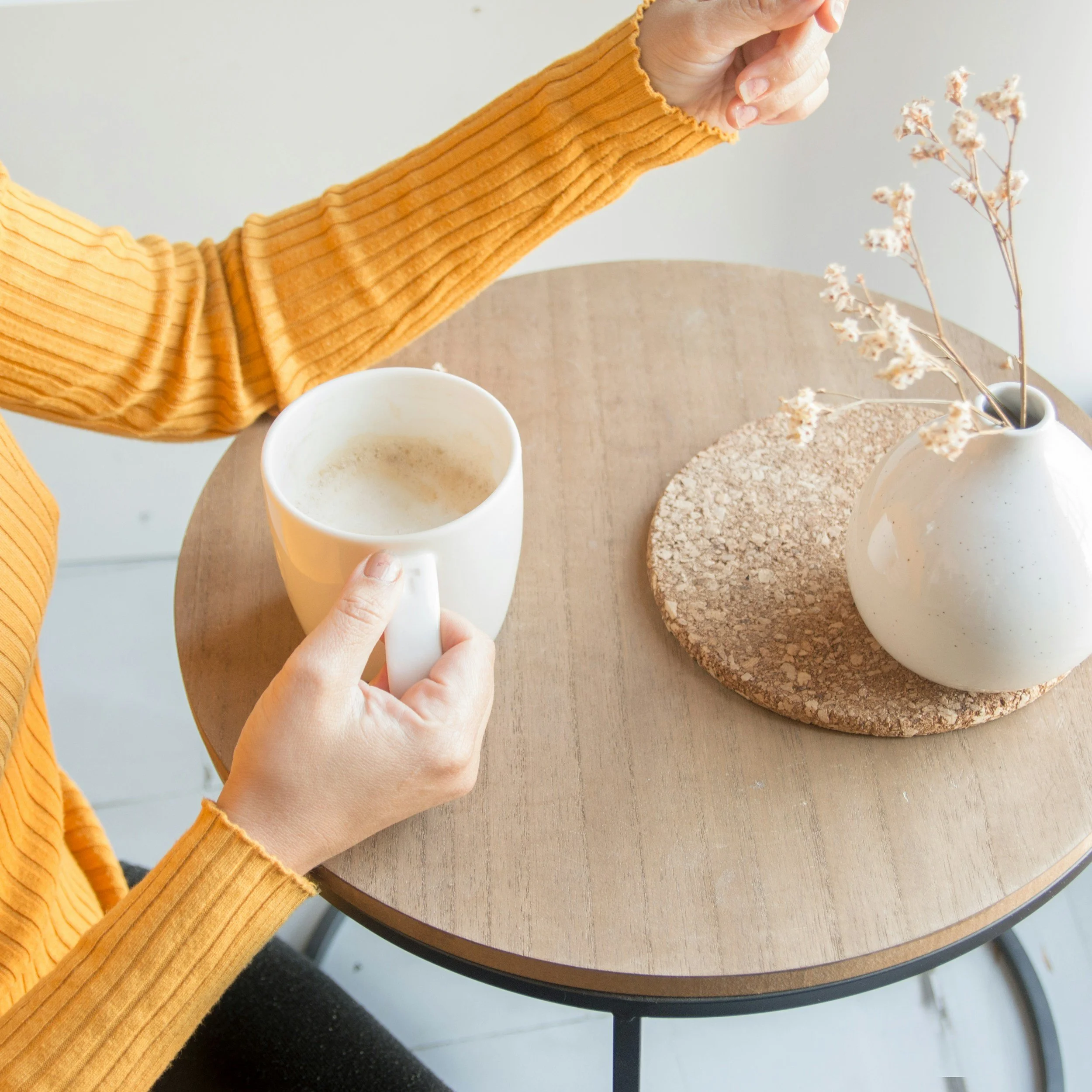 Person holding a white mug filled with a beverage, sitting at a round wooden table with a white vase and dried flowers on a cork coaster.
