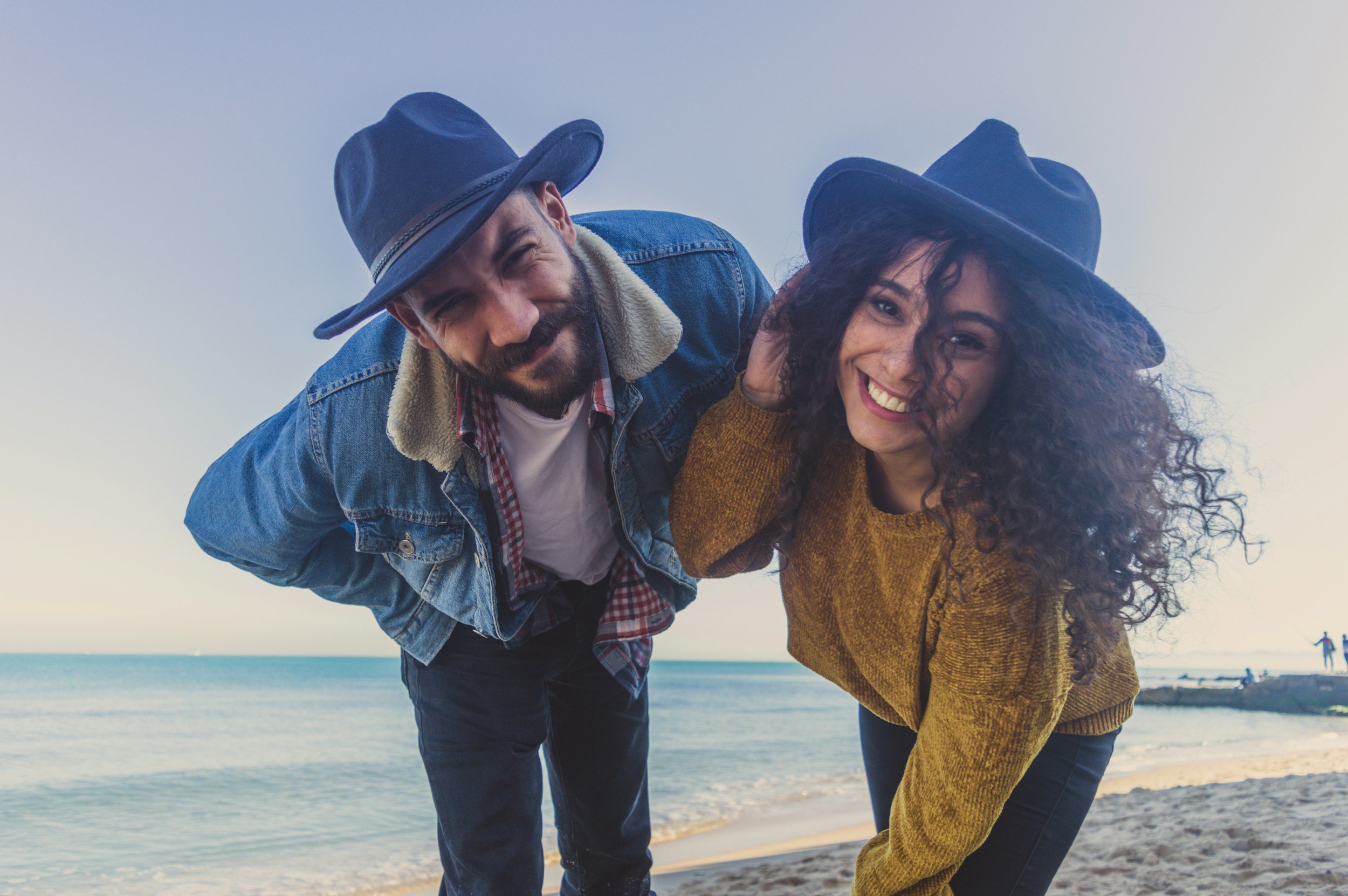 A man and woman on a beach, both wearing black wide-brimmed hats and smiling at the camera. The man is wearing a denim jacket over a plaid shirt, and the woman is wearing a mustard-colored sweater. They are leaning toward each other with the ocean and sky in the background.