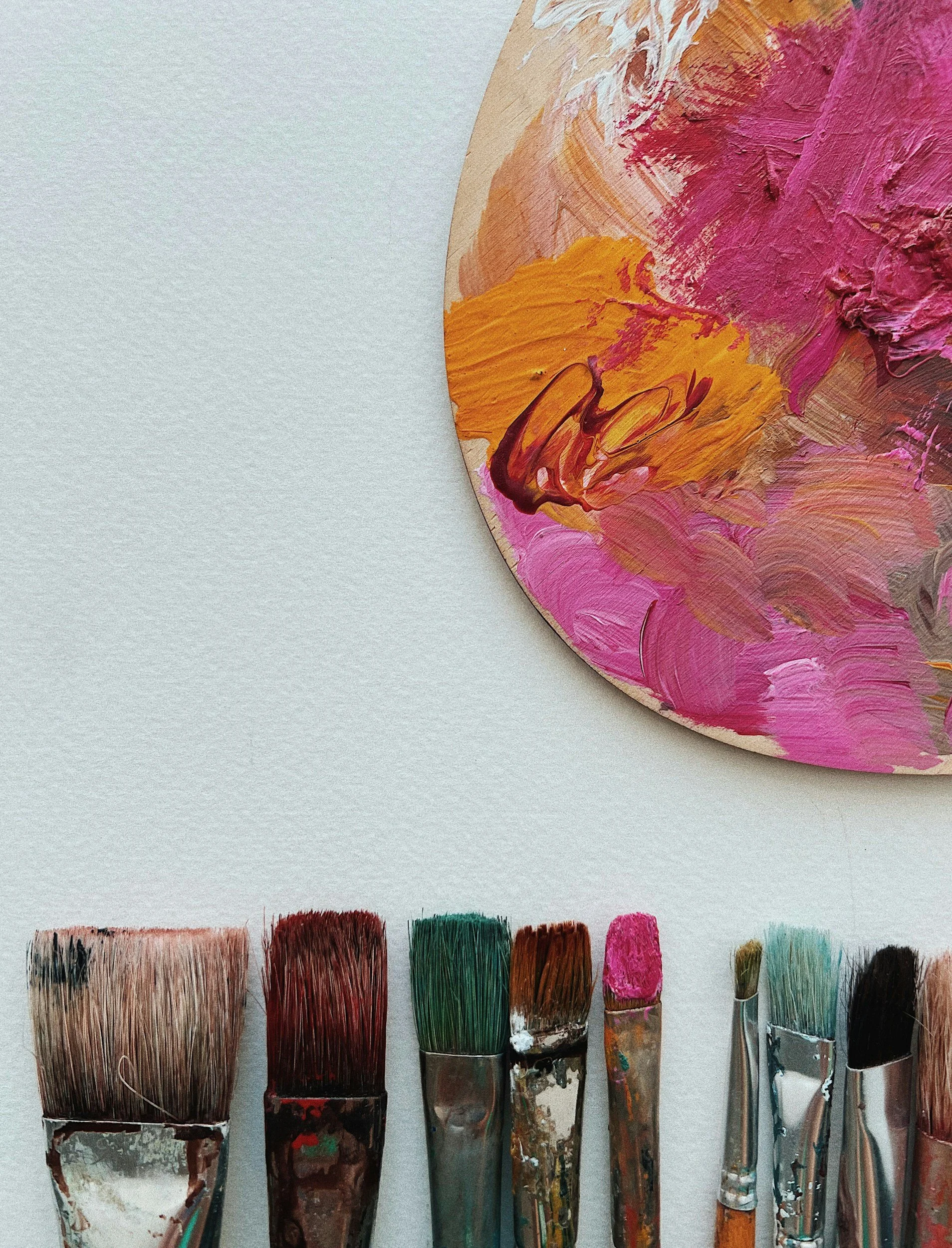 A painter's palette with pink, orange, and white paint next to seven paintbrushes with colorful bristles on a white surface.