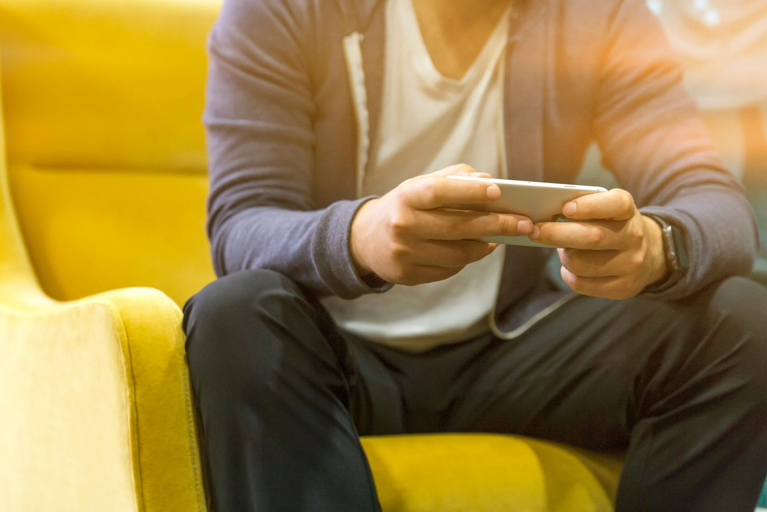 Person sitting on a yellow couch using a smartphone, wearing a gray jacket, white shirt, and black pants.