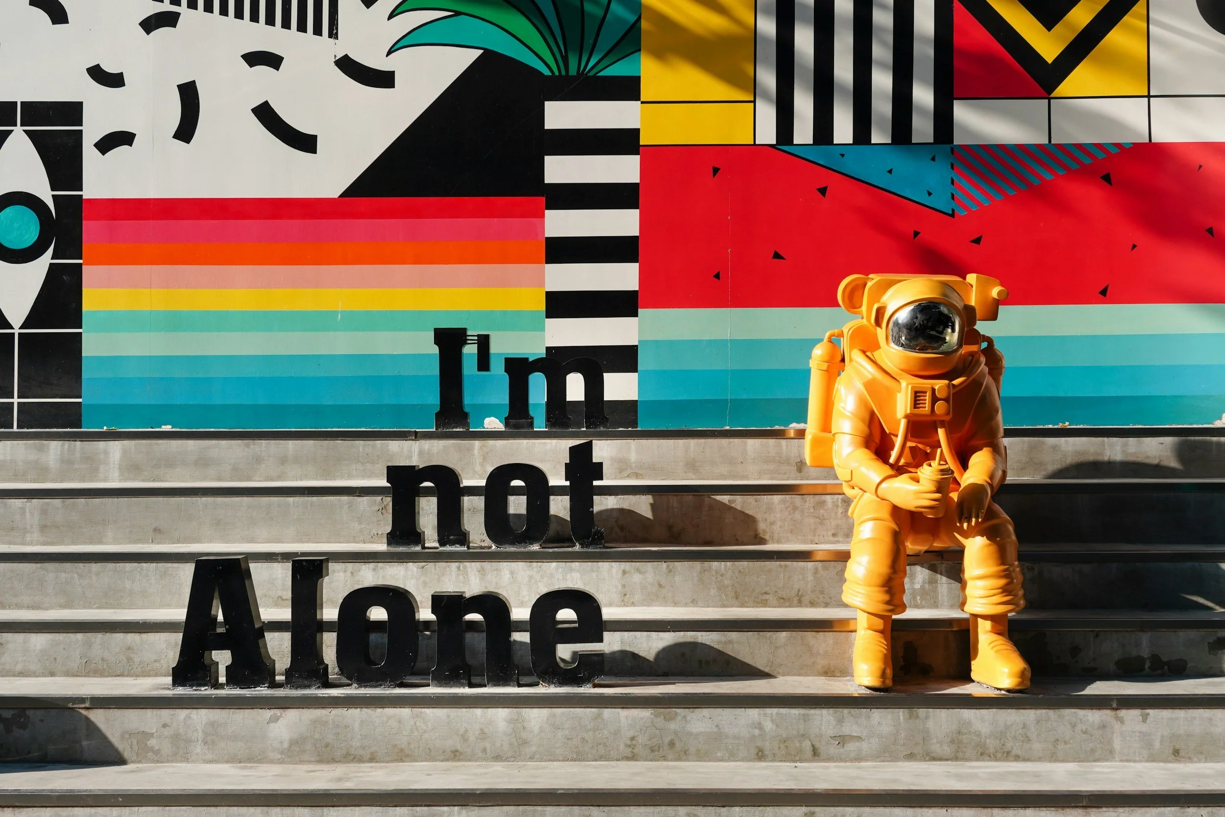 A person in an orange spacesuit sits on steps beside large Im not Alone letters, with a colorful mural behind.
