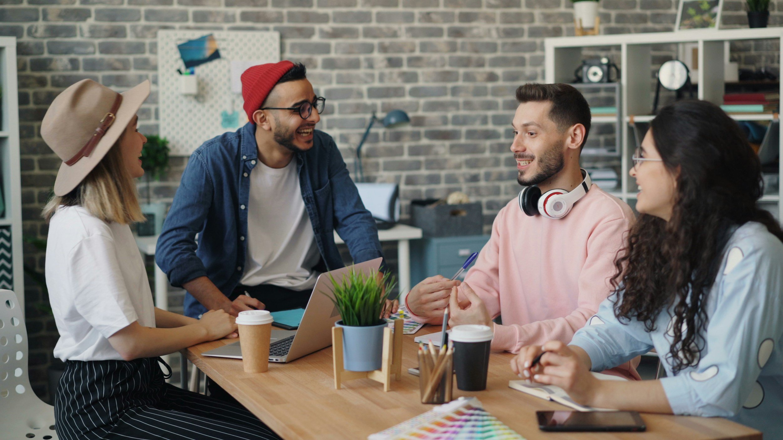 Four young adults sit and laugh around a table with laptops, coffee, and art supplies in a modern, creative office space.