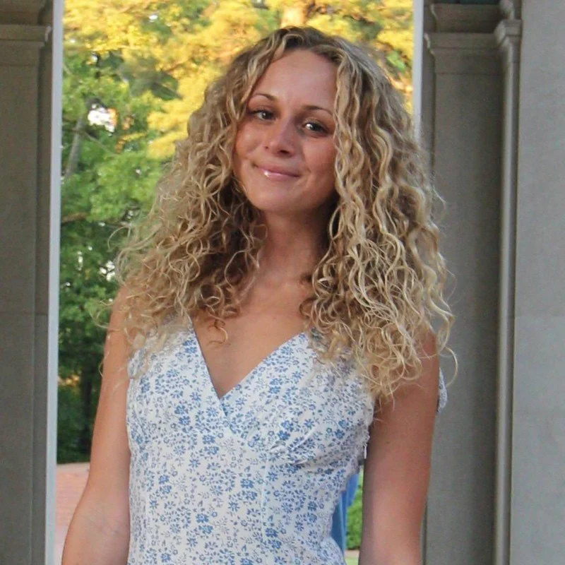 Female therapist with curly blonde hair wearing a sleeveless white and blue floral dress stands outdoors, smiling. Trees are in the background.